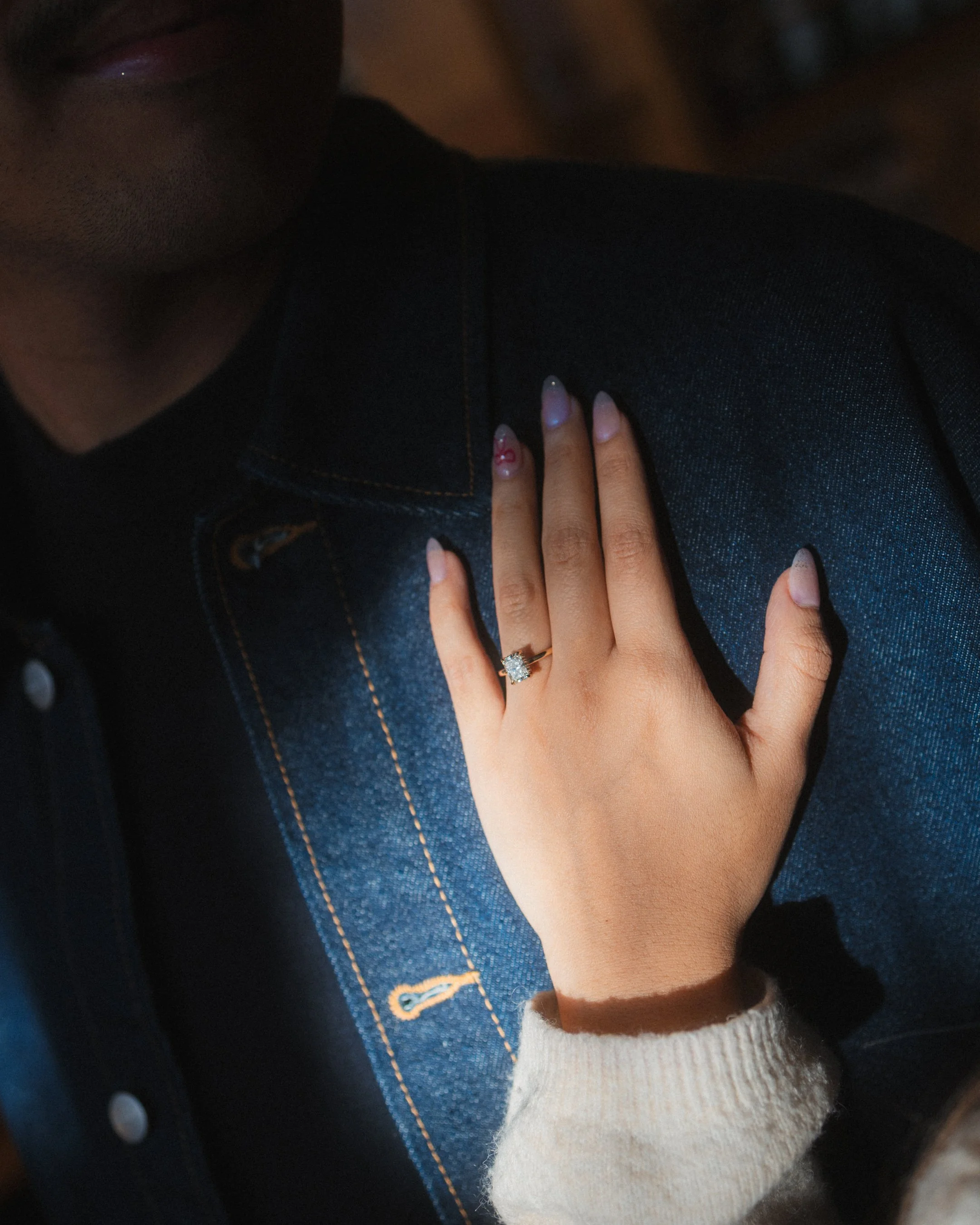 Close-up of a woman's hand with a diamond engagement ring resting on a man's denim jacket.