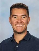 Portrait of a man with short dark hair, wearing a brown button-up shirt, smiling against a blue background.