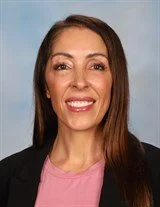 Headshot of a young man with light skin, brown hair, wearing a dark blue collared shirt, smiling against a cloudy sky background.