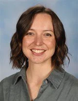 A woman with shoulder-length brown hair, smiling, wearing a dark blouse with small polka dots, against a plain background.