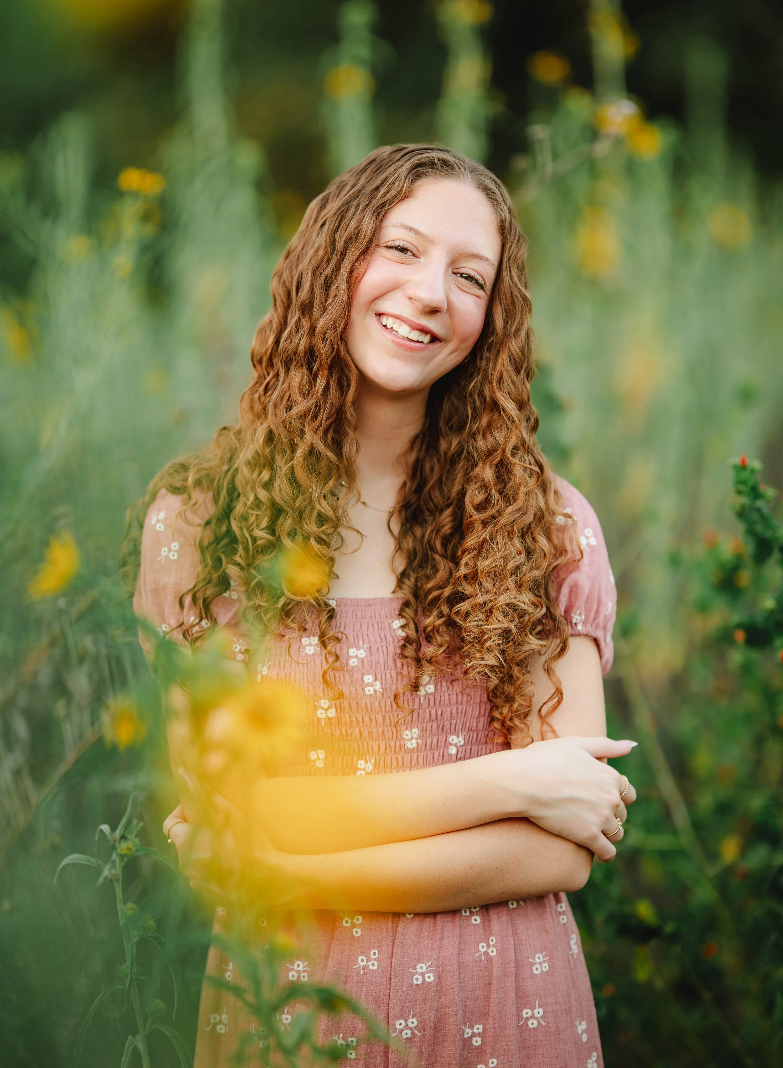 A young woman with curly red hair smiling outdoors in a field of yellow flowers, wearing a pink dress with white floral embroidery.