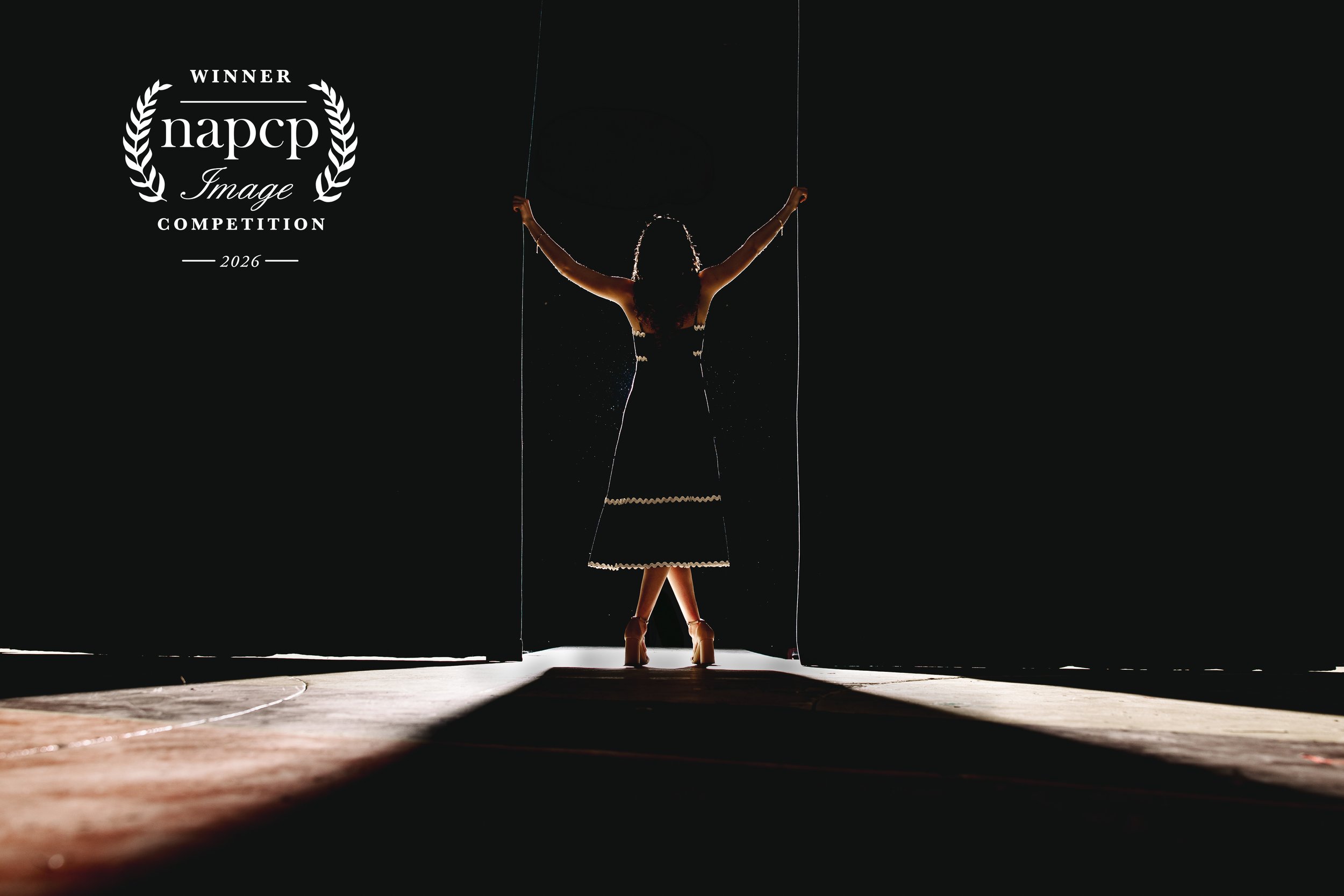 A woman standing behind stage curtains with her arms raised and holding the curtains open, illuminated from behind, creating a silhouette against the dark background. There is a badge that indicates NAPCP winning image.