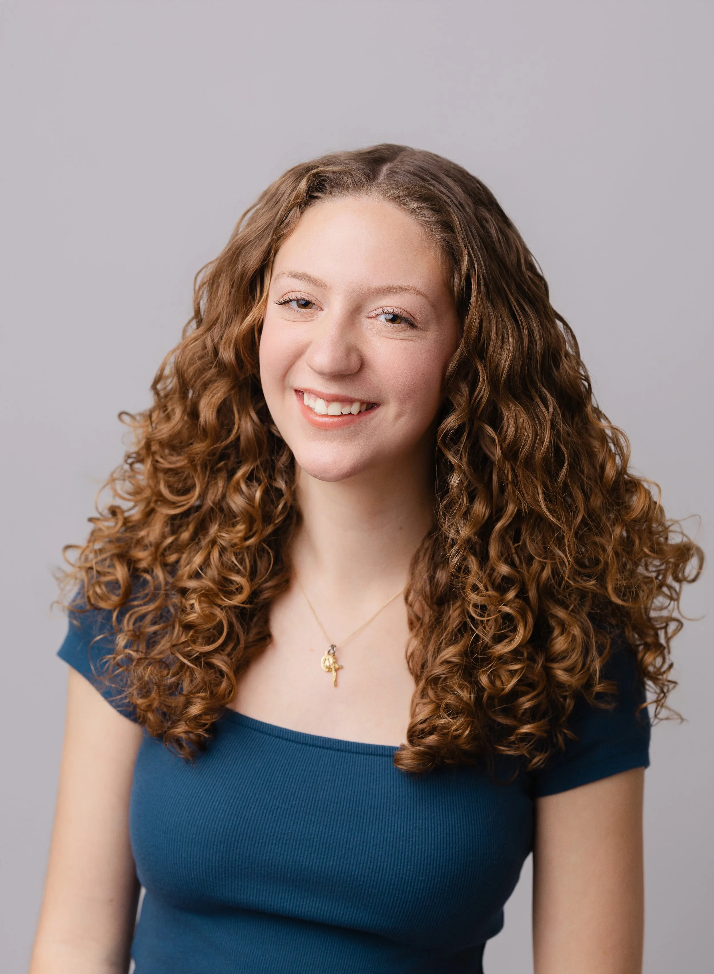 Portrait of a young woman with long, curly brown hair, smiling, wearing a blue top and a gold necklace with a small pendant, against a neutral gray background.