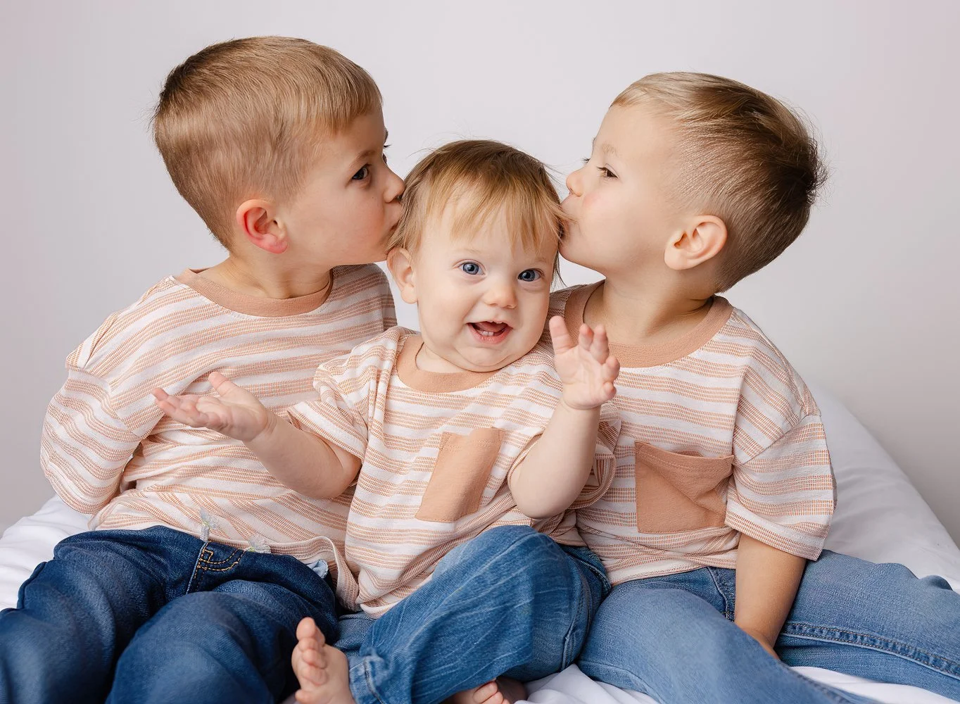 Three young boys wearing matching beige striped t-shirts and blue jeans sitting together on a bed, with two kissing each other on the cheeks and the middle boy smiling with his arms raised.