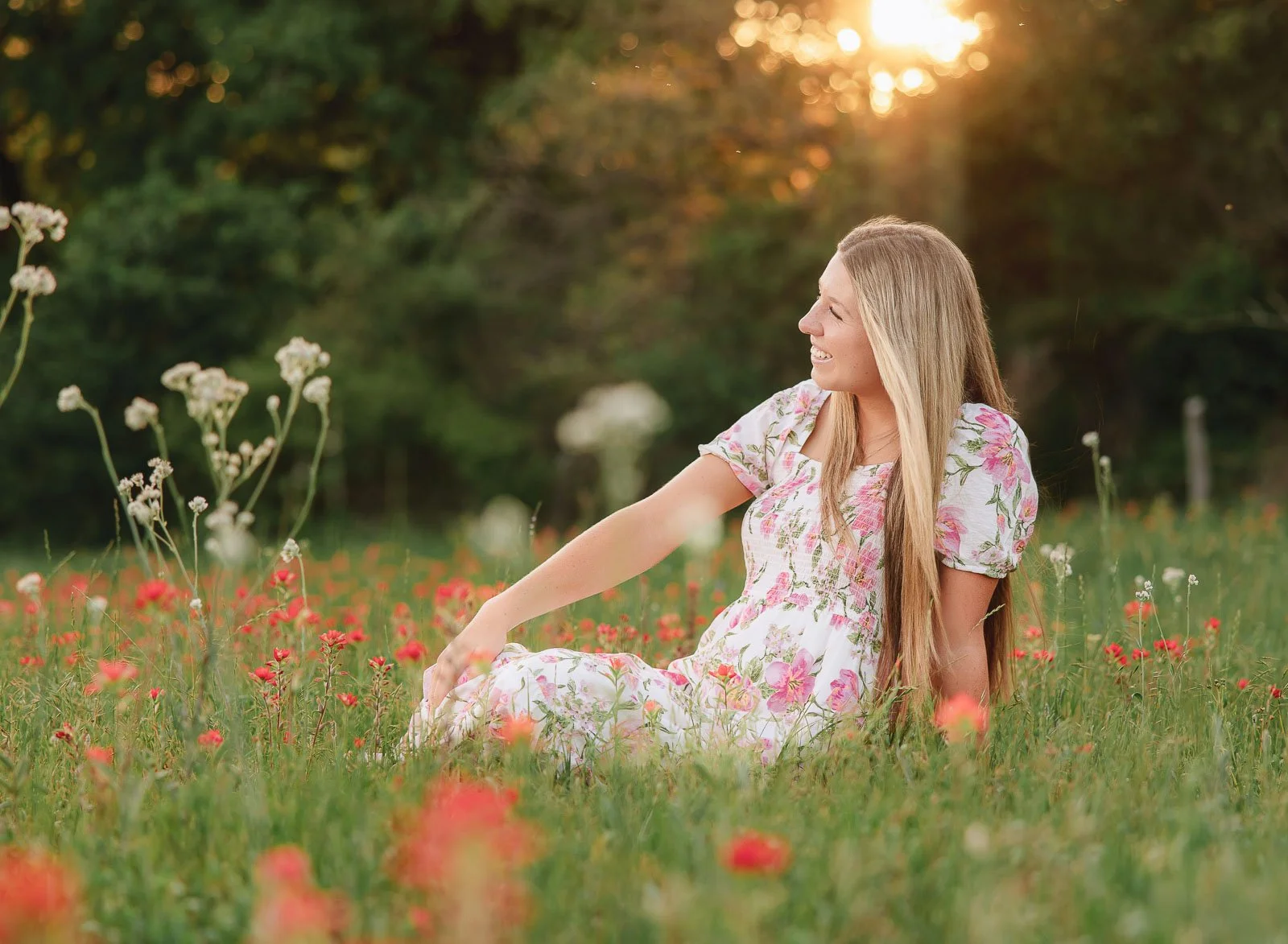 A woman with long blonde hair wearing a floral dress sitting in a field of pink and white flowers during sunset, smiling and looking to the right.