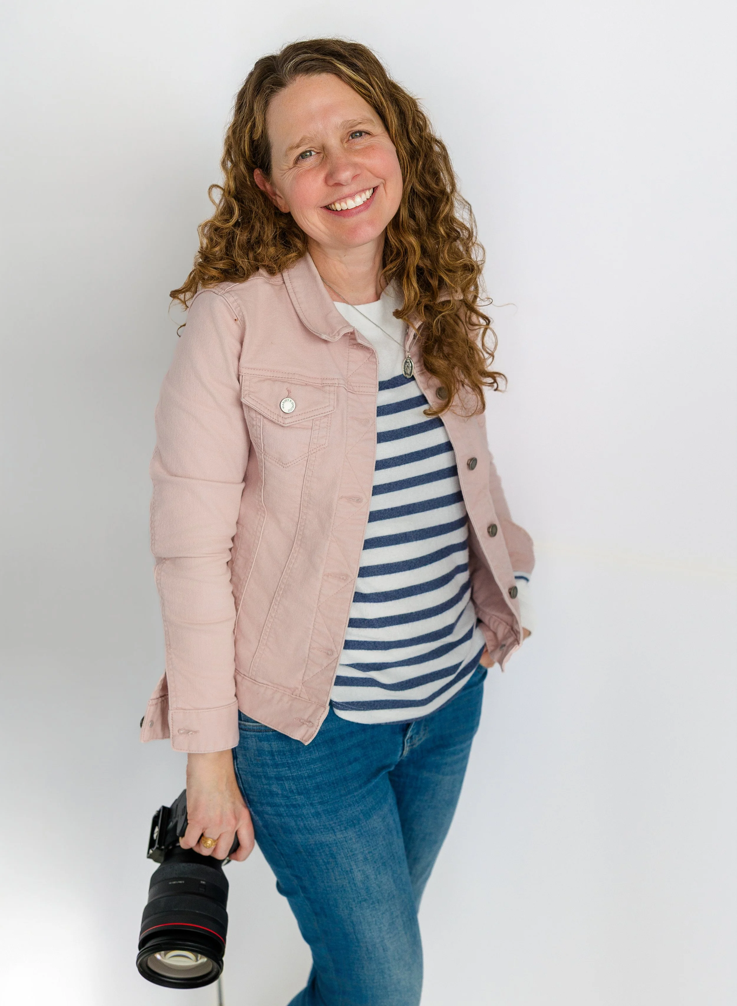 A woman with curly red hair holding a camera in her right hand and smiling at the camera. She is wearing a pink jacket, a striped navy and white shirt, and blue jeans, standing against a plain white background.