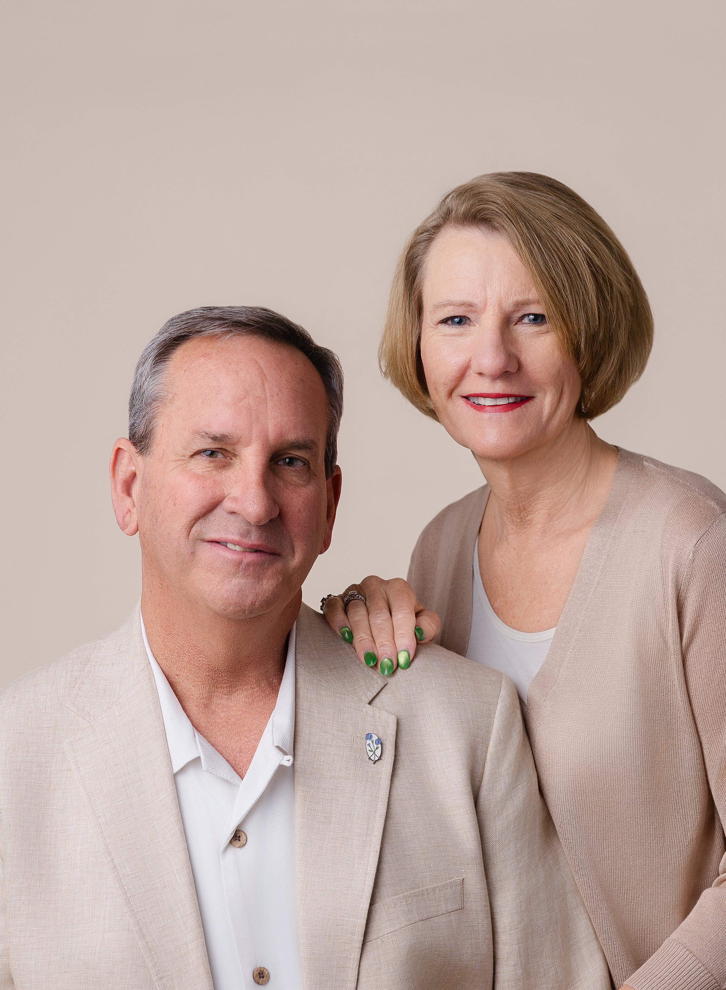 A middle-aged man and woman posing together against a plain background, both dressed in beige suits and smiling at the camera.