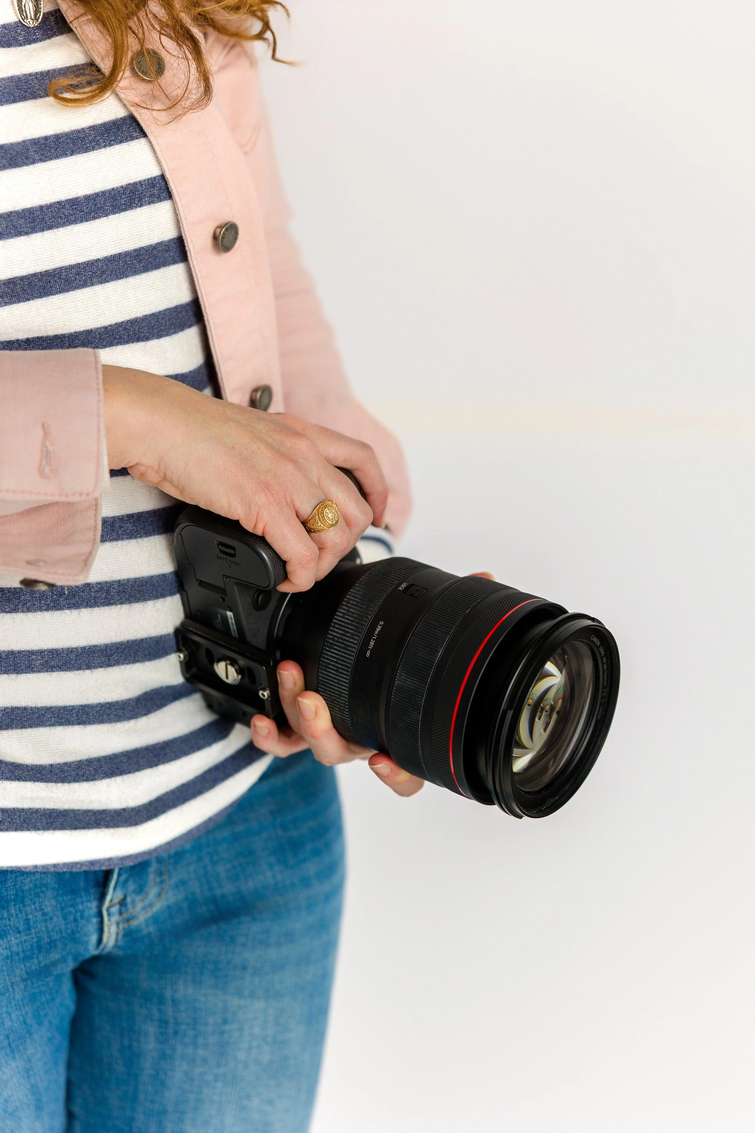 Person wearing a pink jacket and blue jeans holding a professional camera with a large lens, standing against a plain white background.