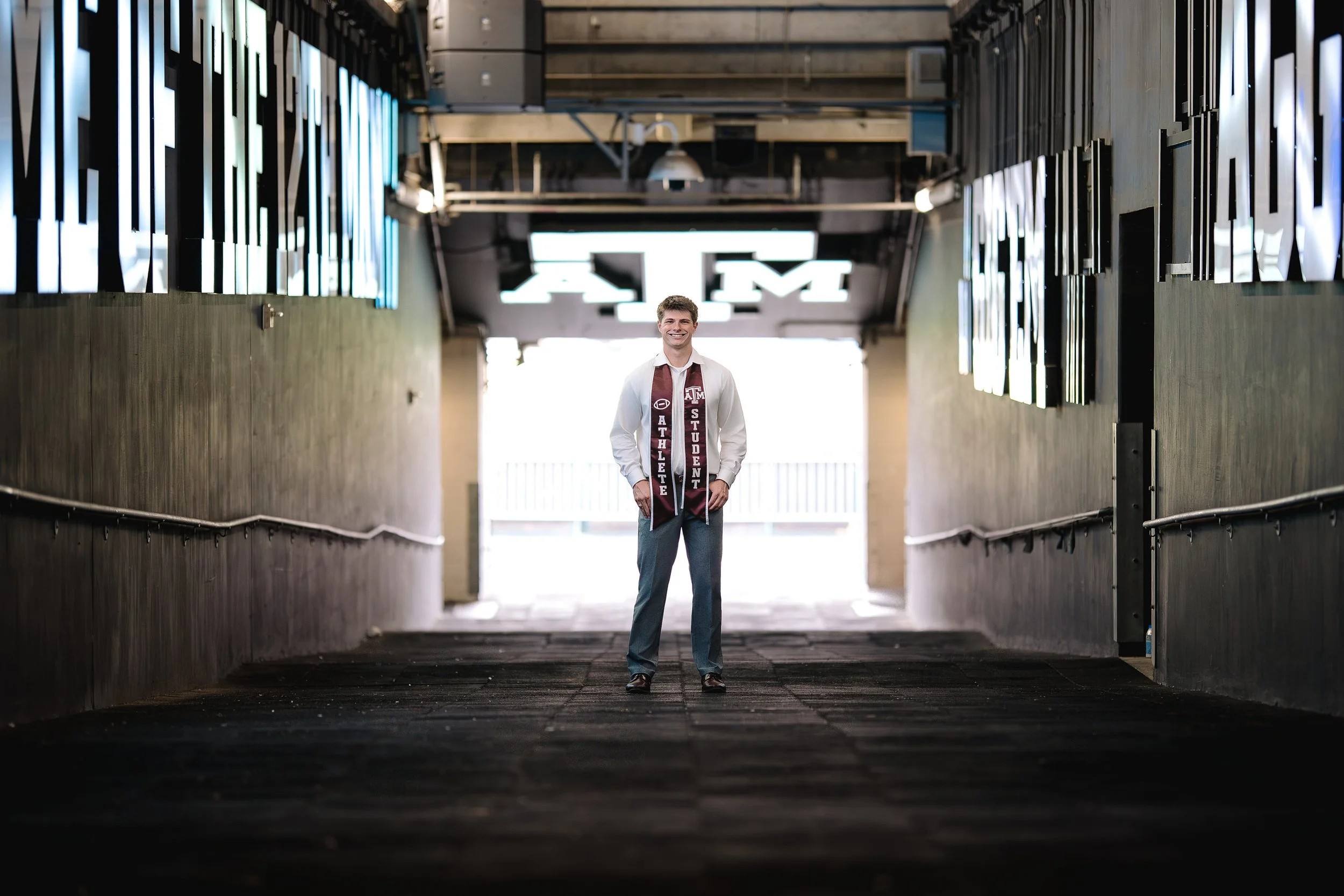 A young man dressed in a white shirt, gray pants, and a maroon scarf with 'TEXAS A&M ATHLETE STUDENT' written on it, walking confidently down a tunnel-like corridor with illuminated signs and the Texas A&M logo overhead, smiling.
