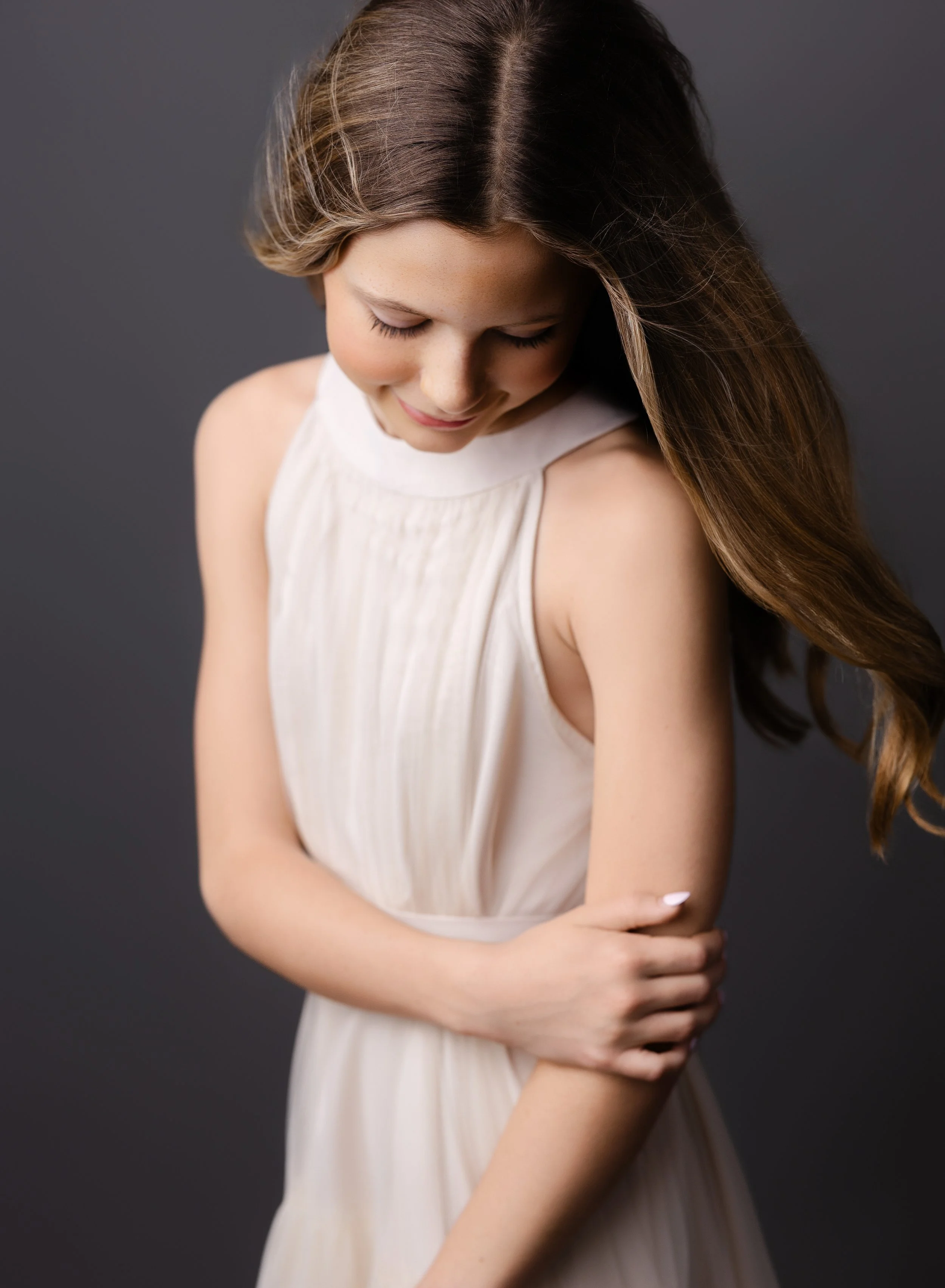 A young girl with long brown hair is smiling and looking down, wearing a sleeveless cream-colored dress, standing against a dark gray background.