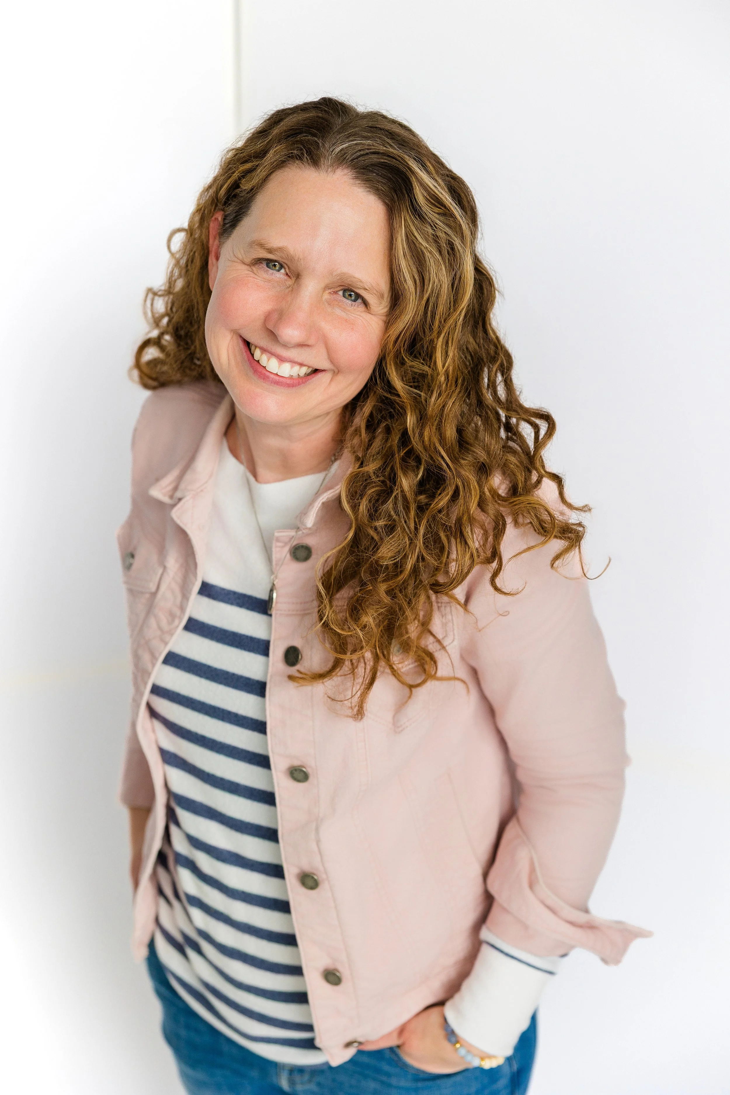 A woman with long curly red hair smiling, wearing a light pink jacket over a white and navy striped shirt, standing against a white background.