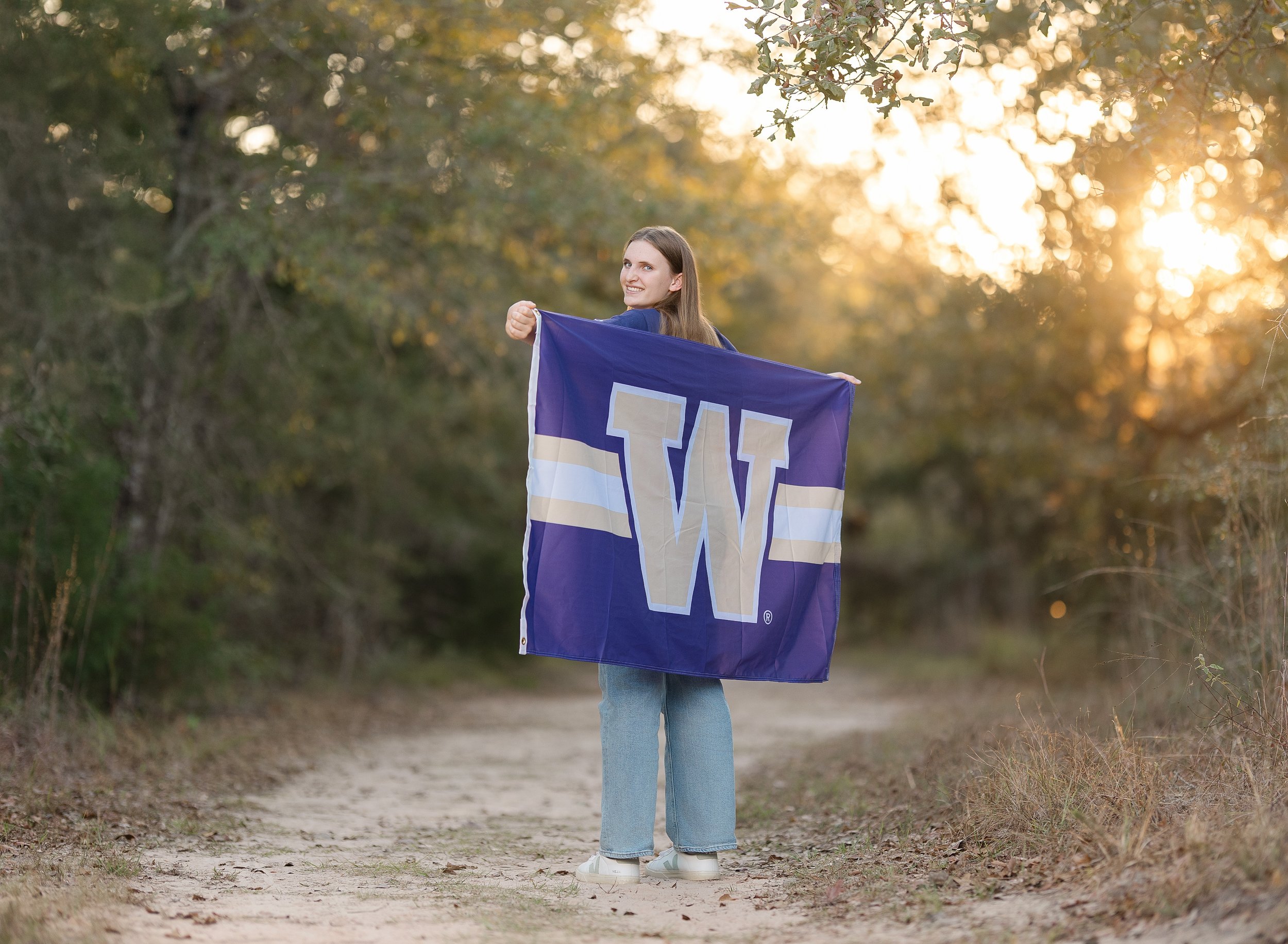 A young woman standing on a dirt path surrounded by trees at sunset, holding a university of Washington flag.