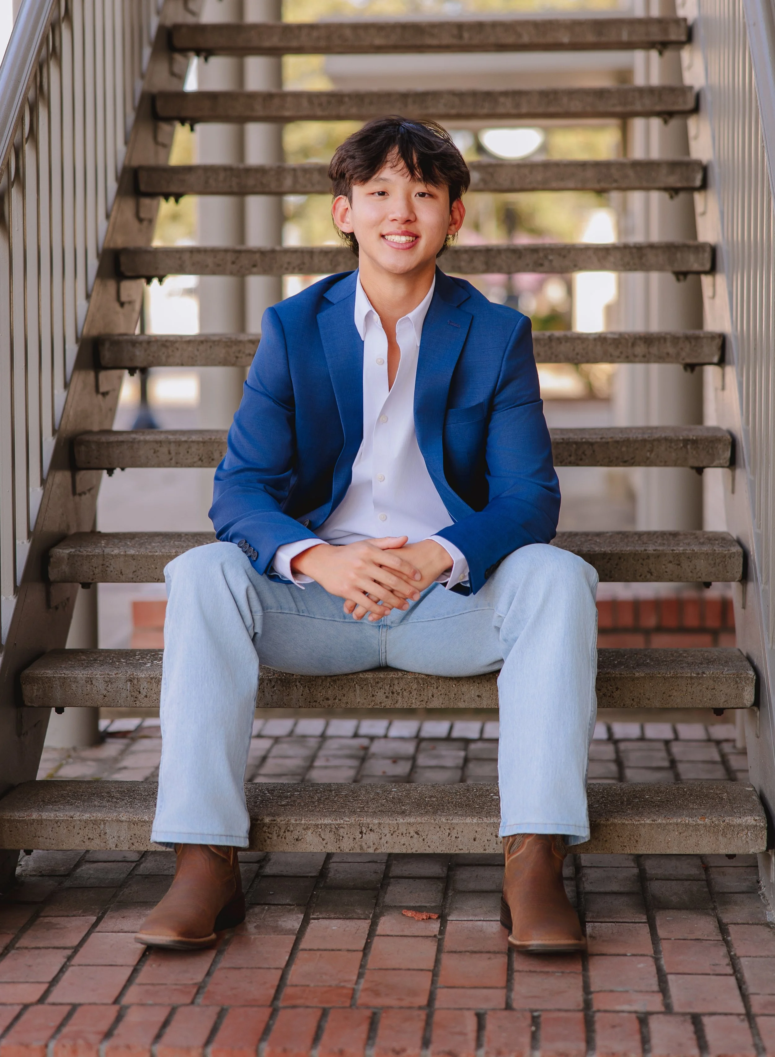 A young man sitting on outdoor stairs, wearing a blue blazer, white shirt, light blue jeans, and brown boots, smiling at the camera.