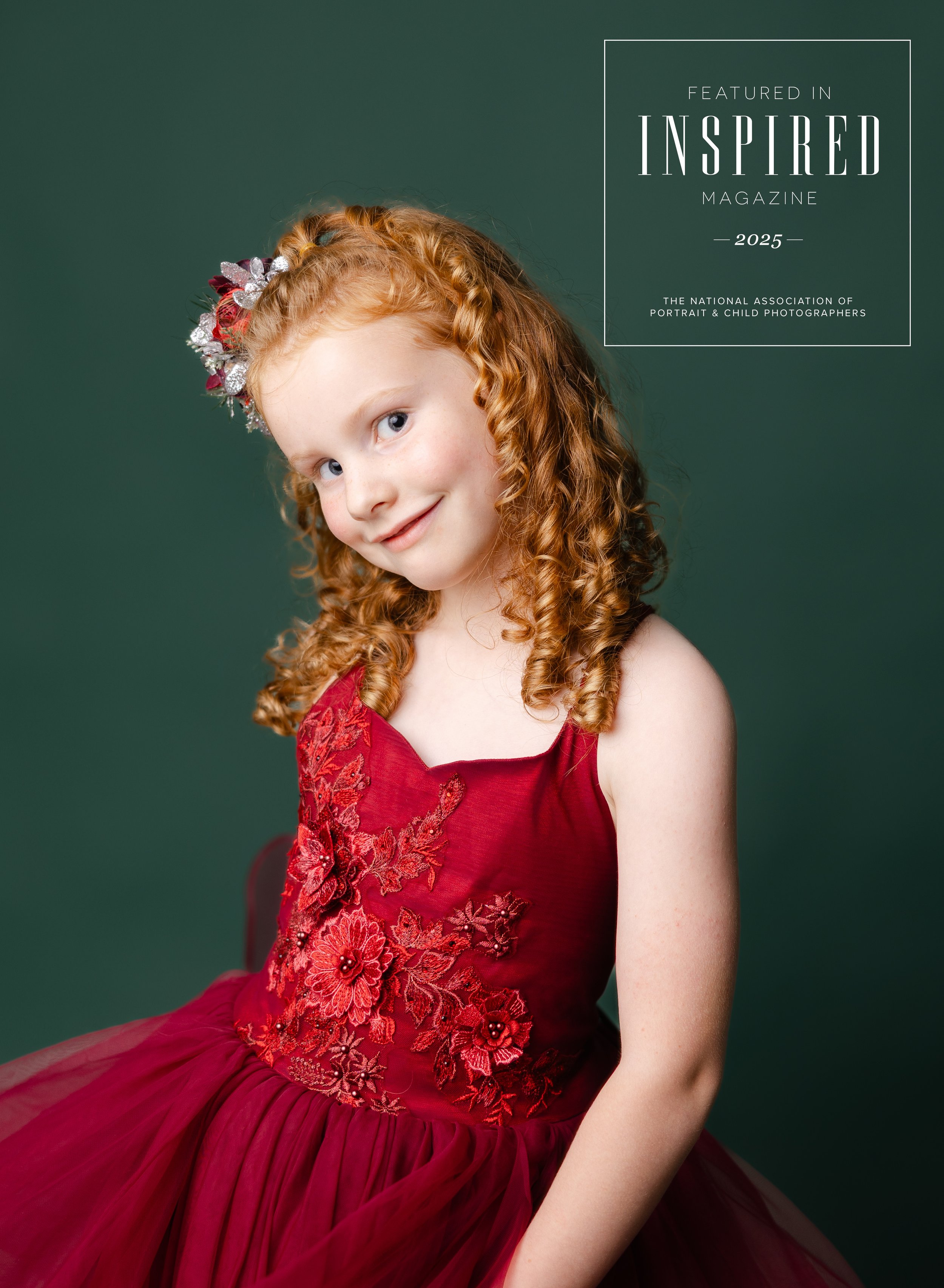 Young girl with curly red hair wearing a red dress with floral embroidery, smiling at the camera, against a dark green background.