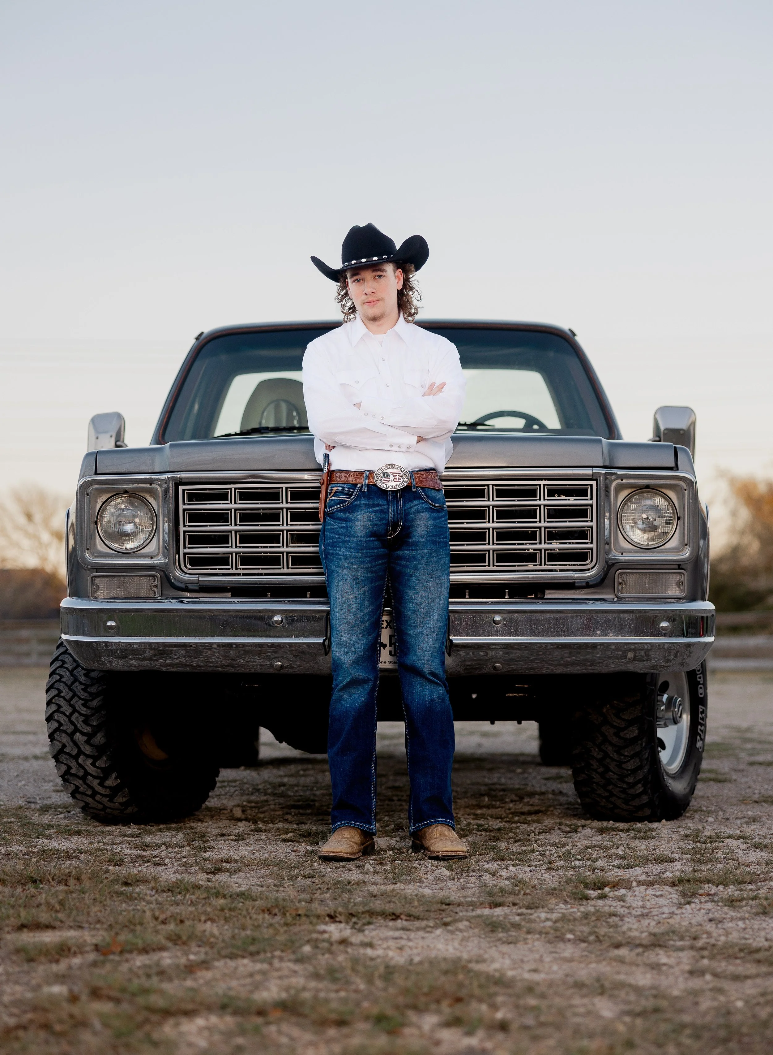 A young man in a white shirt, blue jeans, and cowboy hat standing with arms crossed in front of a black pickup truck outdoors during sunset.