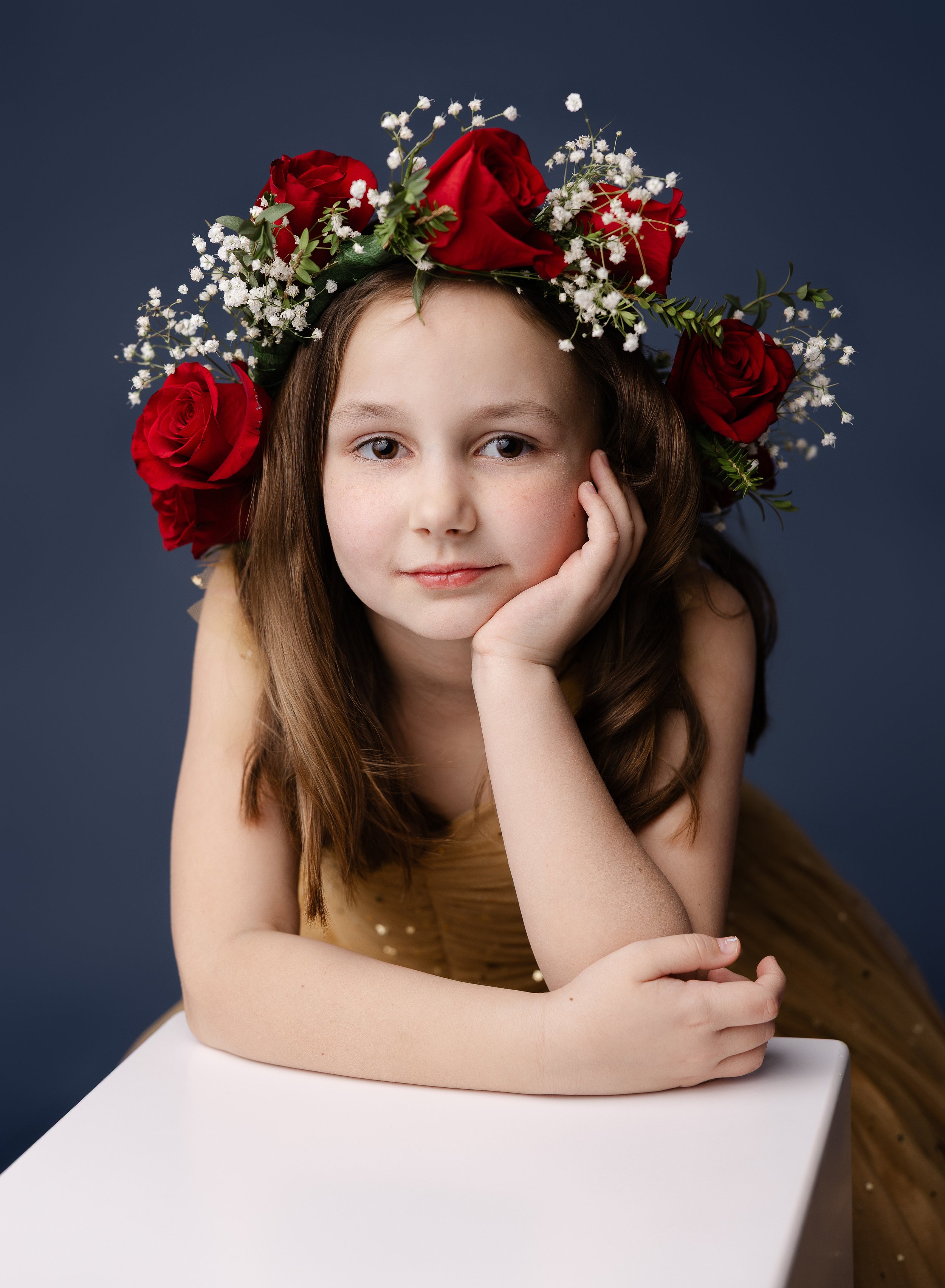 A young girl with long brown hair resting her chin on her left hand, wearing a floral crown of red roses and white baby's breath, and a tan dress with small pearls, against a dark blue background.