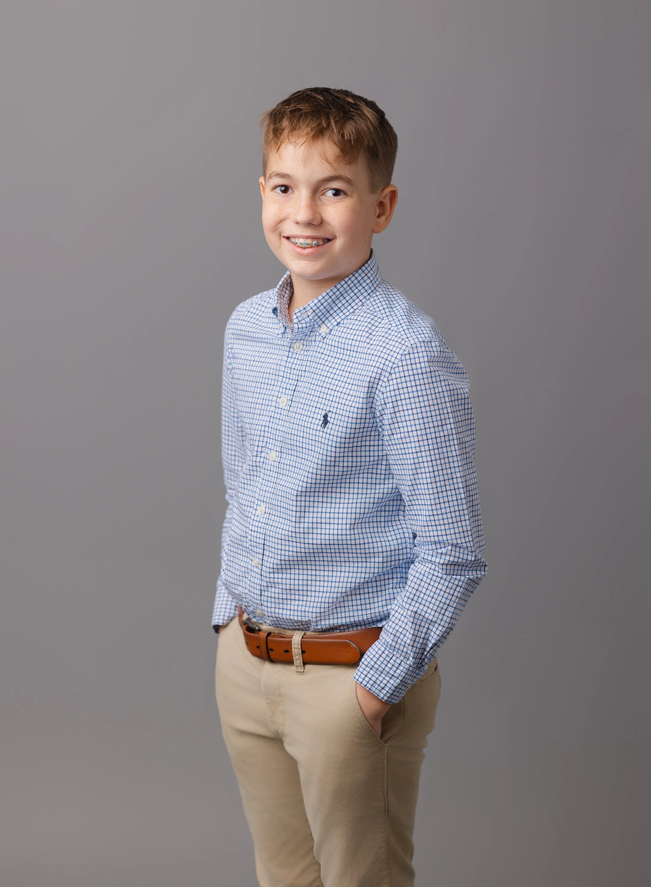 A teenage boy with brown hair, smiling, wearing a light blue checkered button-up shirt, tan pants, and a brown belt, standing against a gray background.
