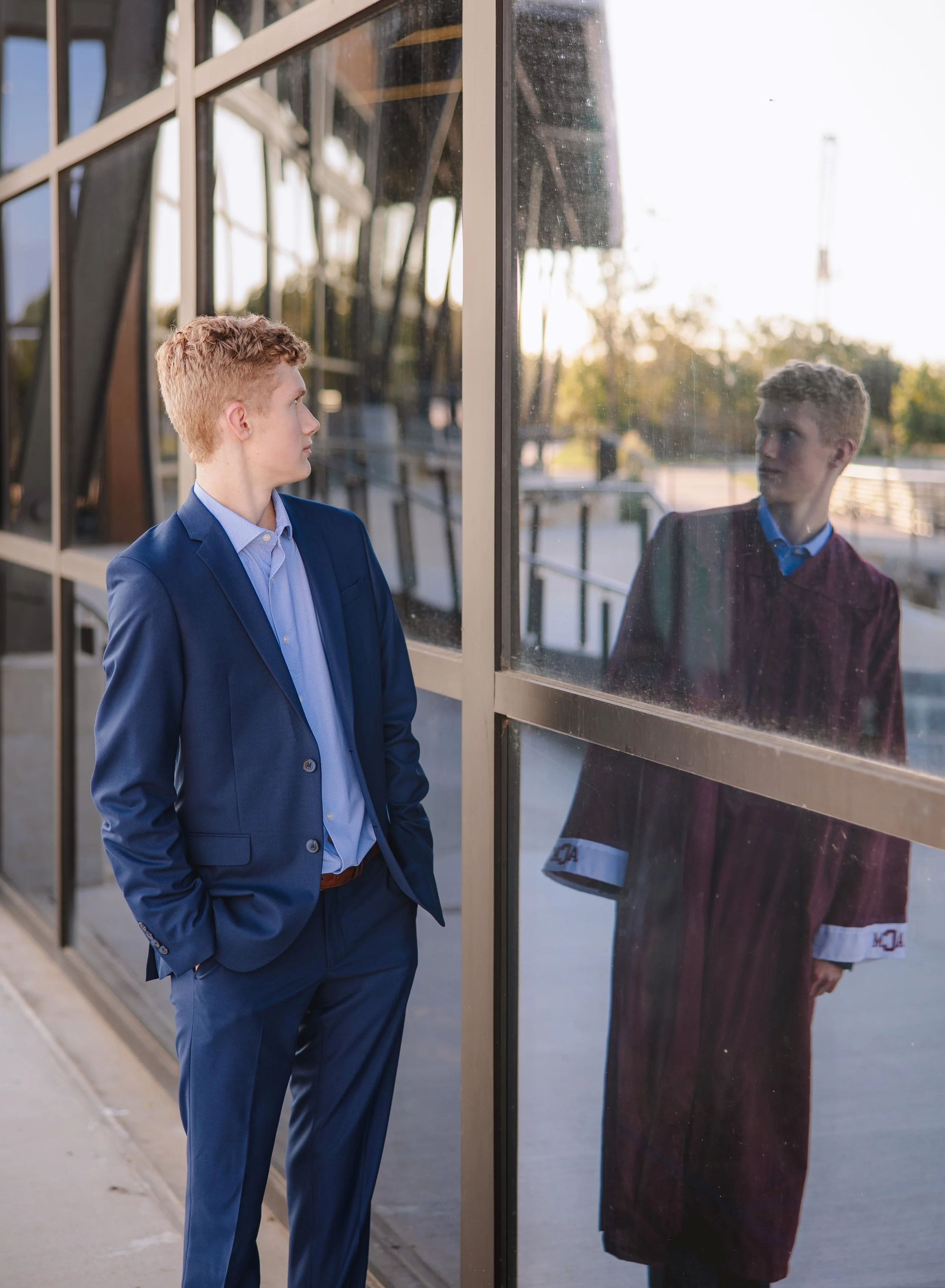 A young man in a blue suit and dress shirt looks at his reflection in a glass window outside a modern building during sunset.