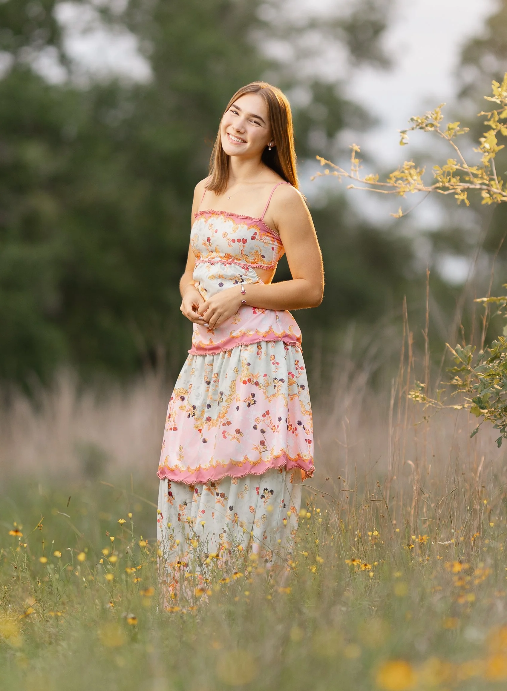Young woman in a floral dress smiling and standing outdoors in a grassy field during sunset.