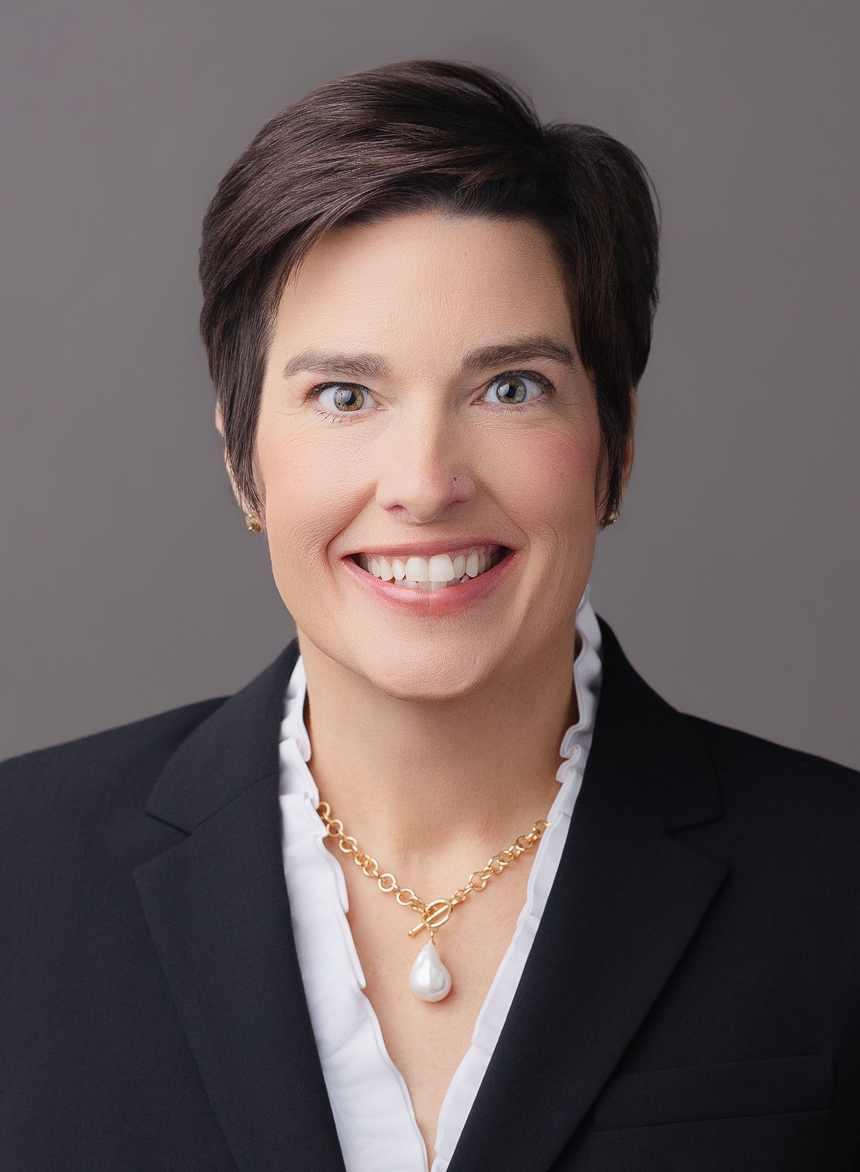Professional headshot of a woman with short dark hair, blue eyes, wearing a black blazer, white shirt with ruffled collar, gold necklace with a large pearl pendant, smiling against a gray background.
