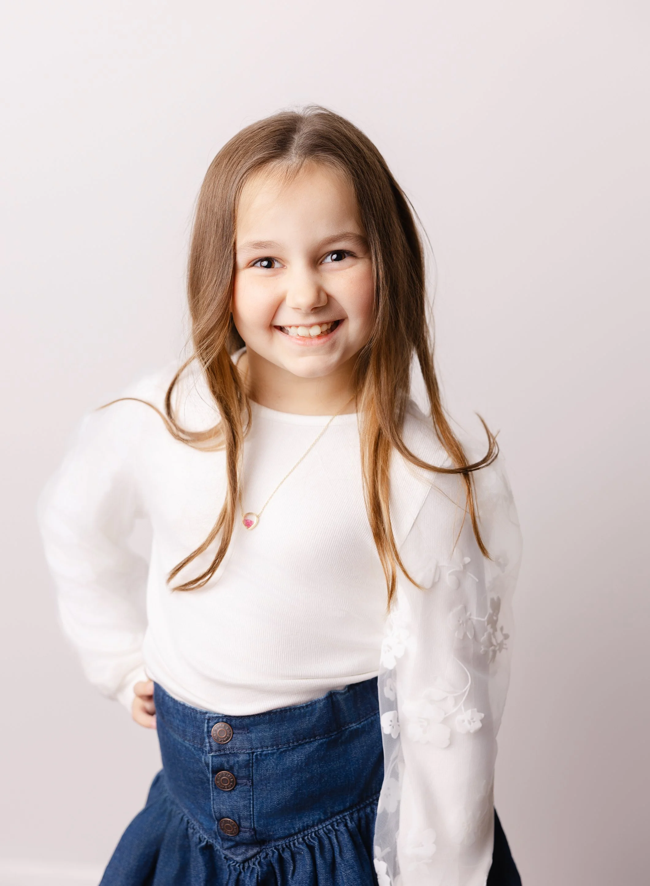 A young girl with long brown hair, smiling, wearing a white long-sleeve top with floral embroidery sleeves, a dark blue skirt with buttons, and a necklace with a pink and gold pendant, standing against a plain white background.