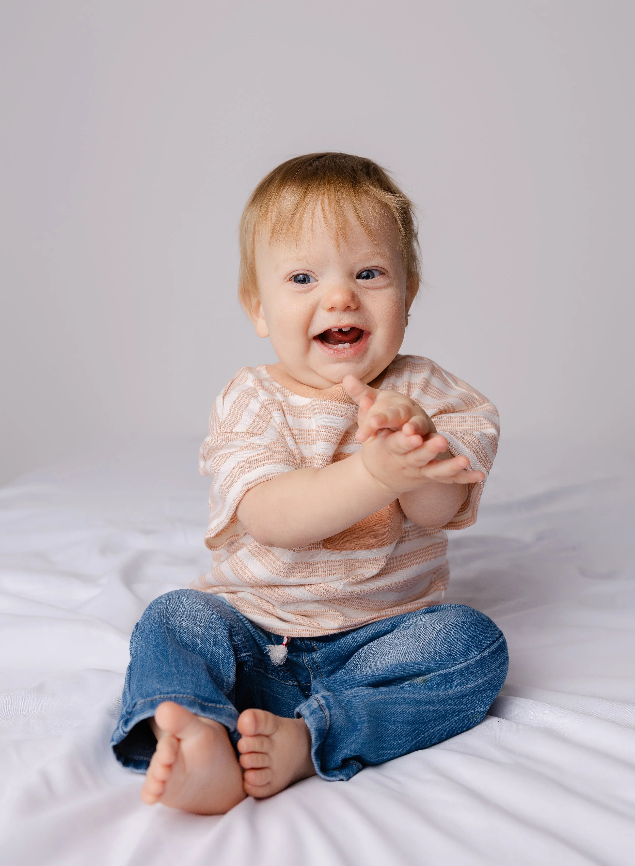 Smiling baby with light brown hair clapping hands, sitting on white bedsheet, wearing a striped beige t-shirt and blue jeans, against a plain gray background.