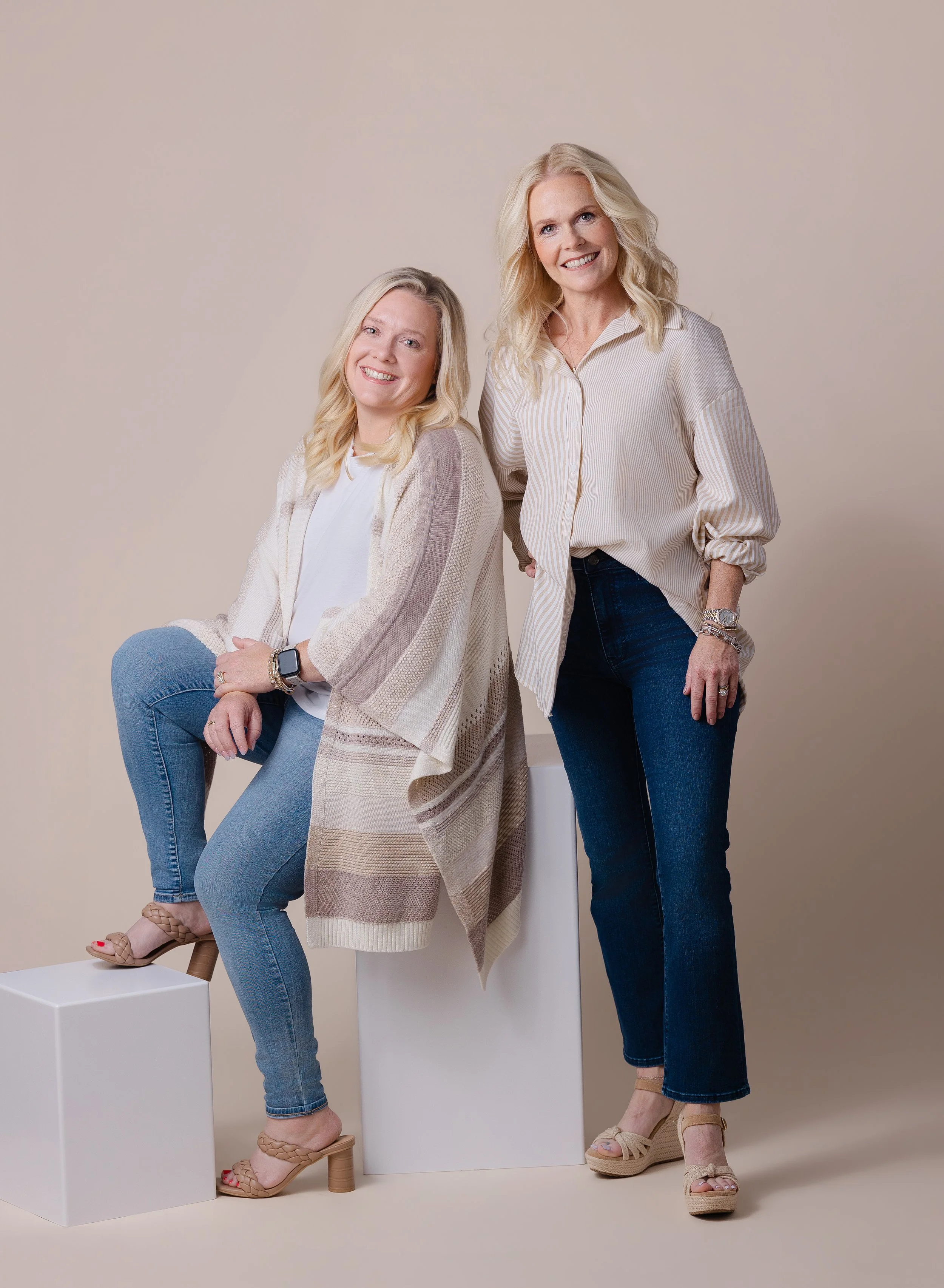 Two women with blonde hair smiling, one seated on a white block with one leg crossed, the other standing with her hand on her hip, both wearing casual beige and white clothing and heels, against a plain beige background.
