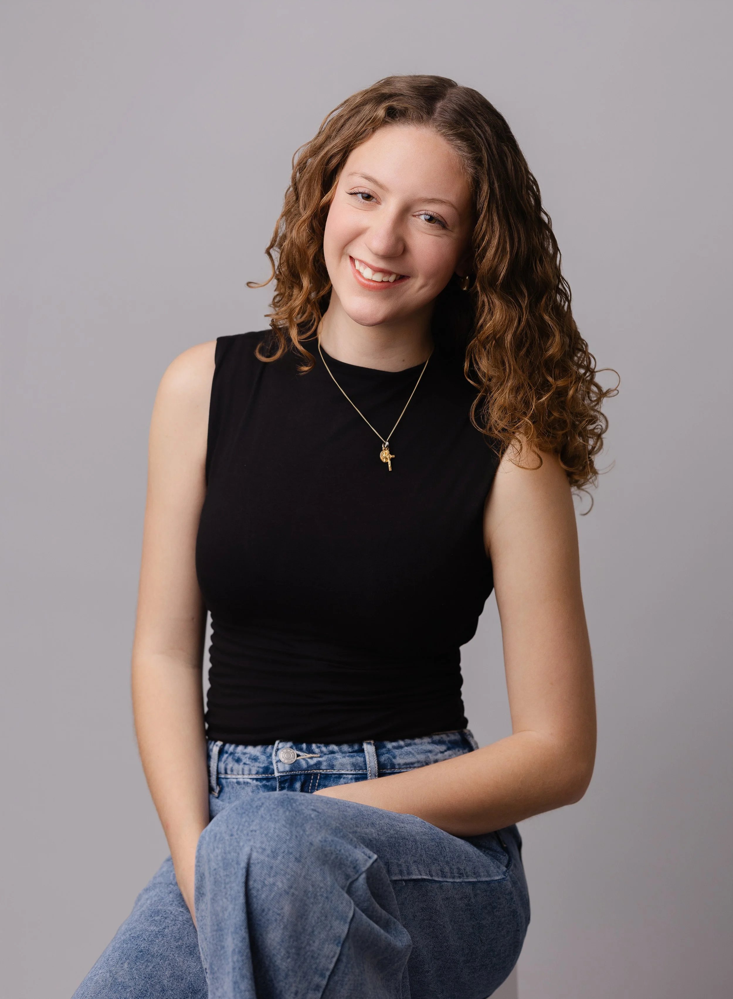 A young woman with curly brown hair wearing a black sleeveless top, blue jeans, and a gold necklace with a cross pendant, smiling against a plain gray background.