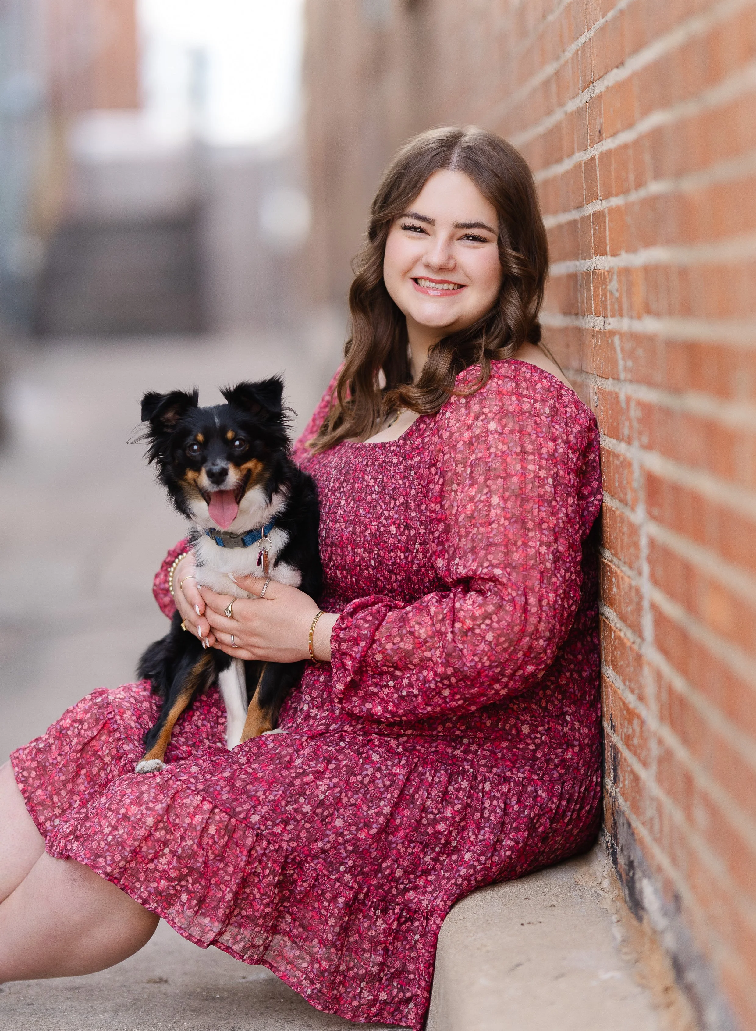 A young woman in a pink floral dress sitting against a brick wall, holding a small black and tan dog with a blue collar, both smiling.