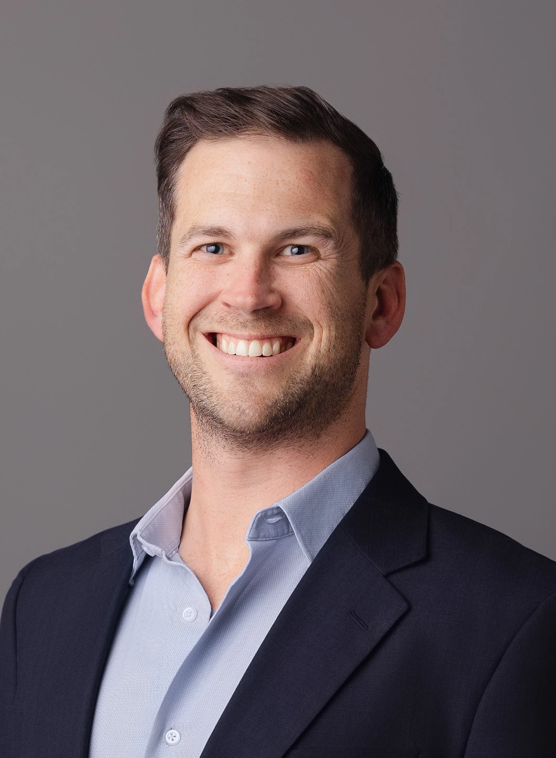 Headshot of a man with short brown hair, blue eyes, wearing a dark suit jacket and light blue collared shirt, smiling against a gray background.