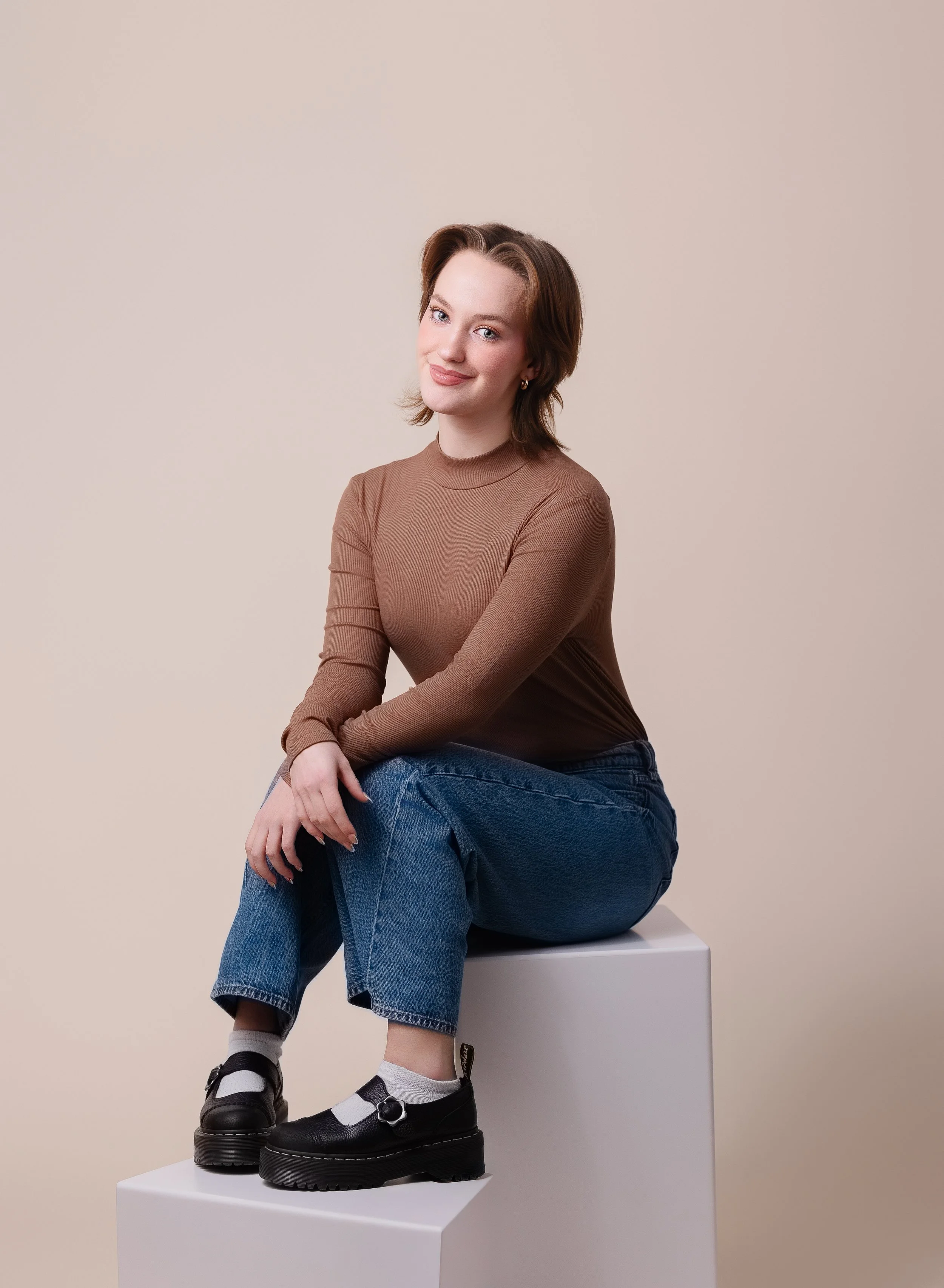 A young woman with brown hair and fair skin sitting on a white cube against a beige background, wearing a brown long-sleeve shirt, blue jeans, and black shoes with socks.