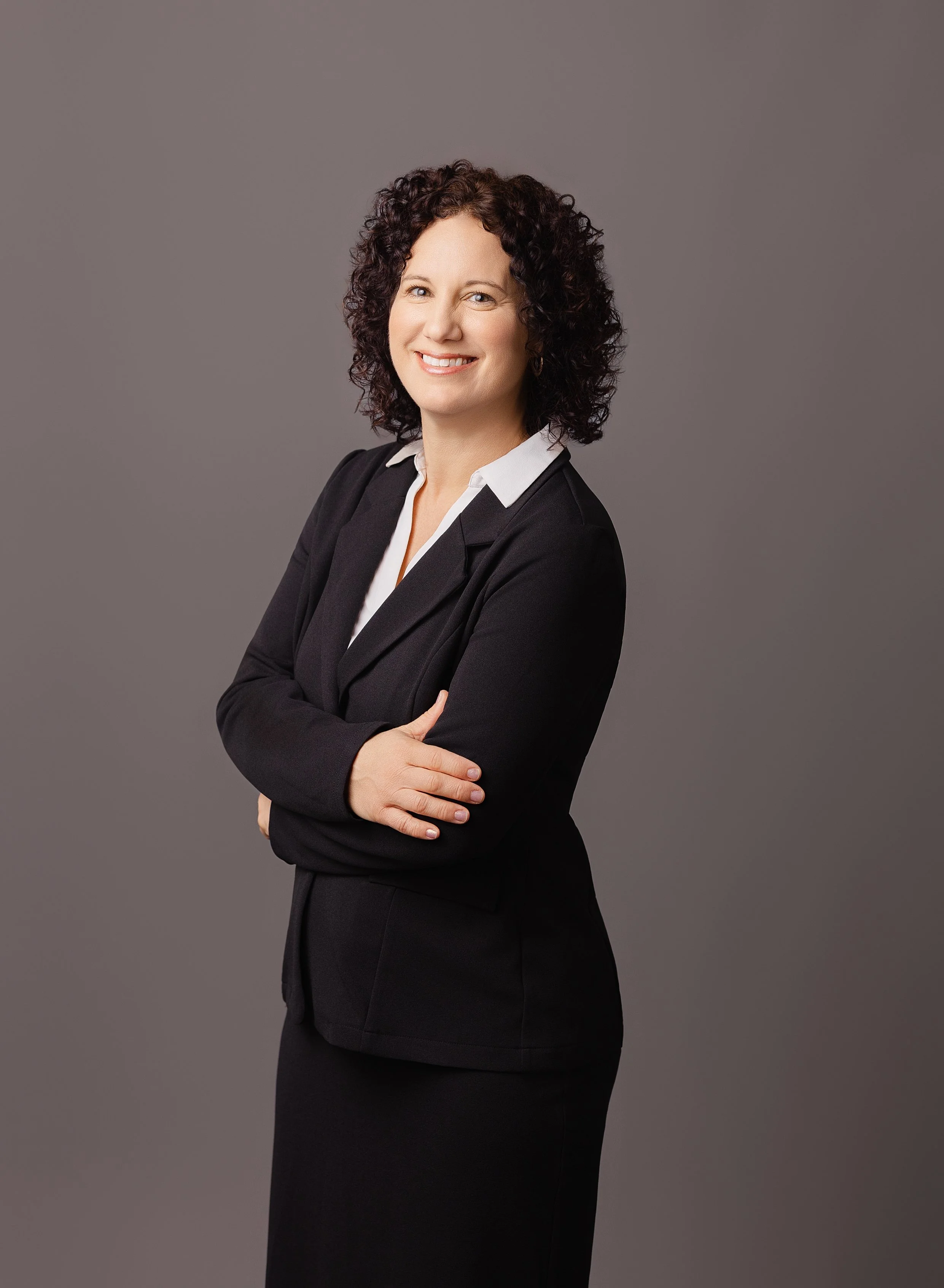 A woman with curly dark hair wearing a black blazer and white blouse, smiling with arms crossed, against a gray background.