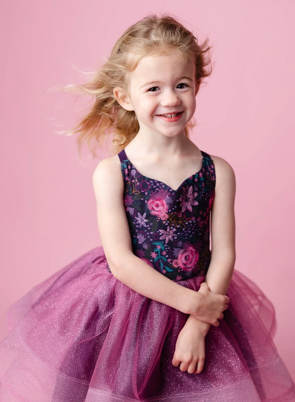 A young girl with blonde hair, smiling, wearing a floral top and a pink tulle dress, standing against a pink background.