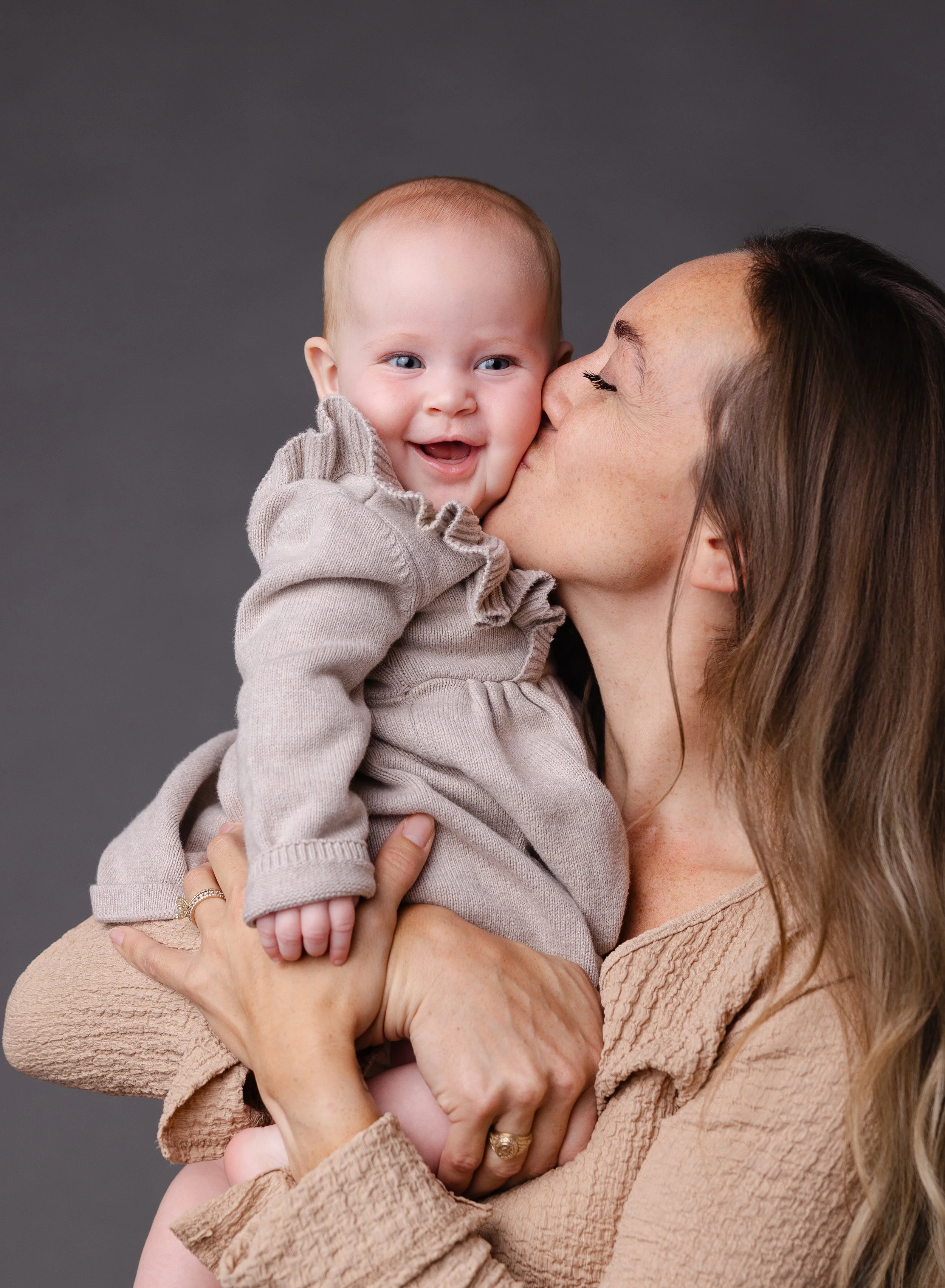 A woman with long brown hair holding a smiling baby with blue eyes and light hair, against a dark gray background.
