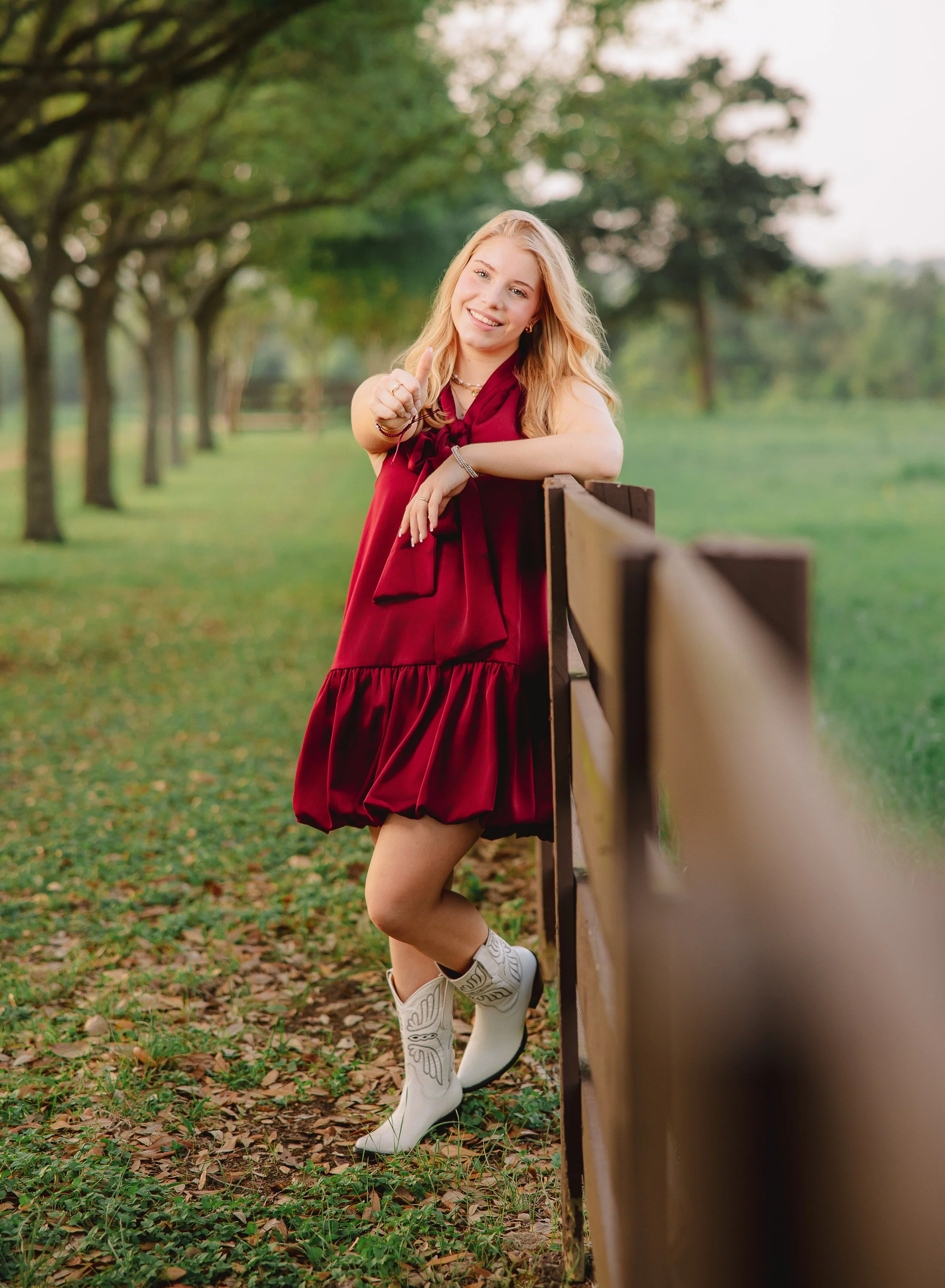 A young woman with blonde hair in a red dress and white cowboy boots standing outdoors near a wooden fence in a green park with trees.