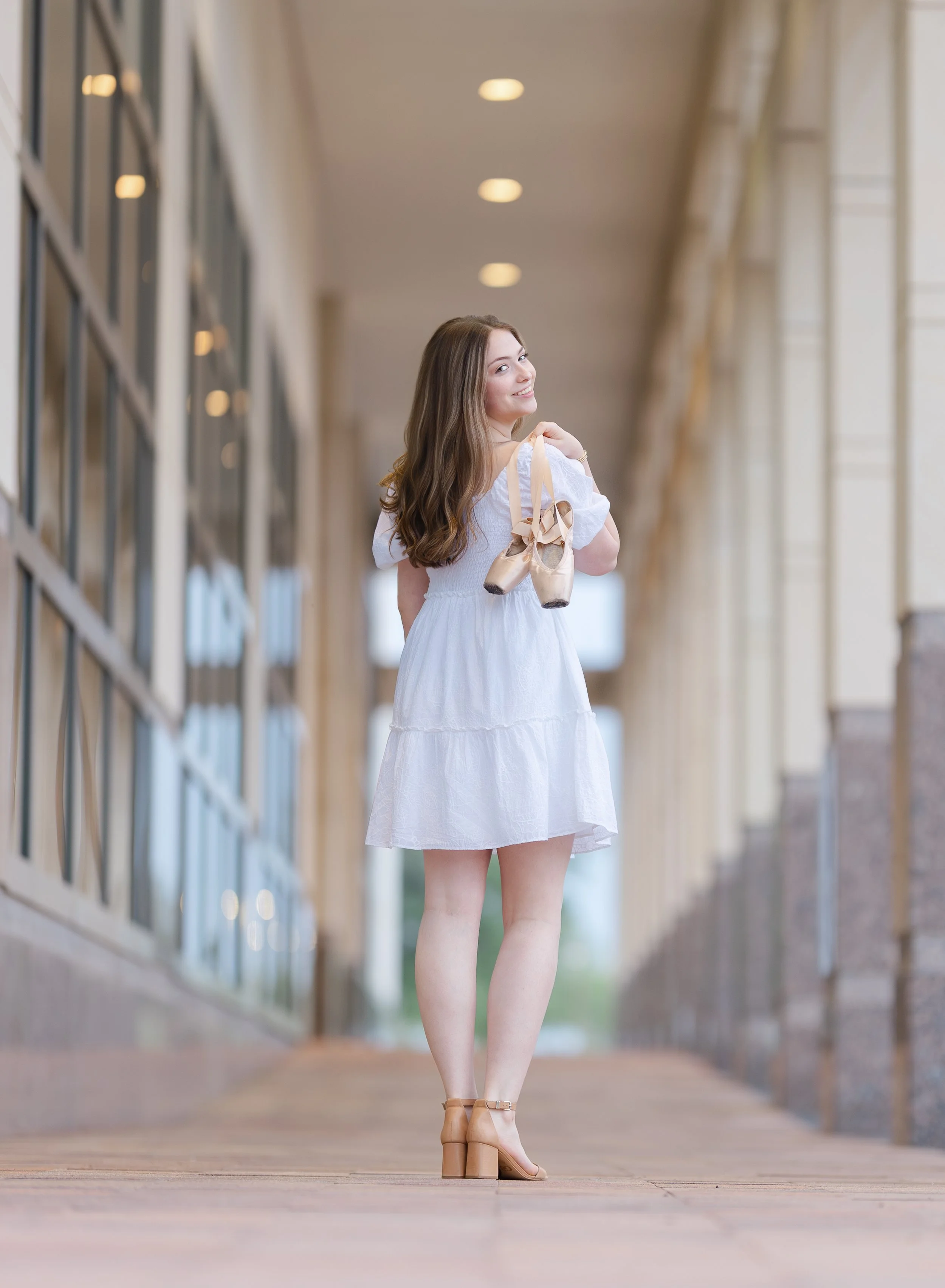 A young woman in a white dress and beige sandals walking through a corridor holding ballet shoes over her shoulder, smiling at the camera.