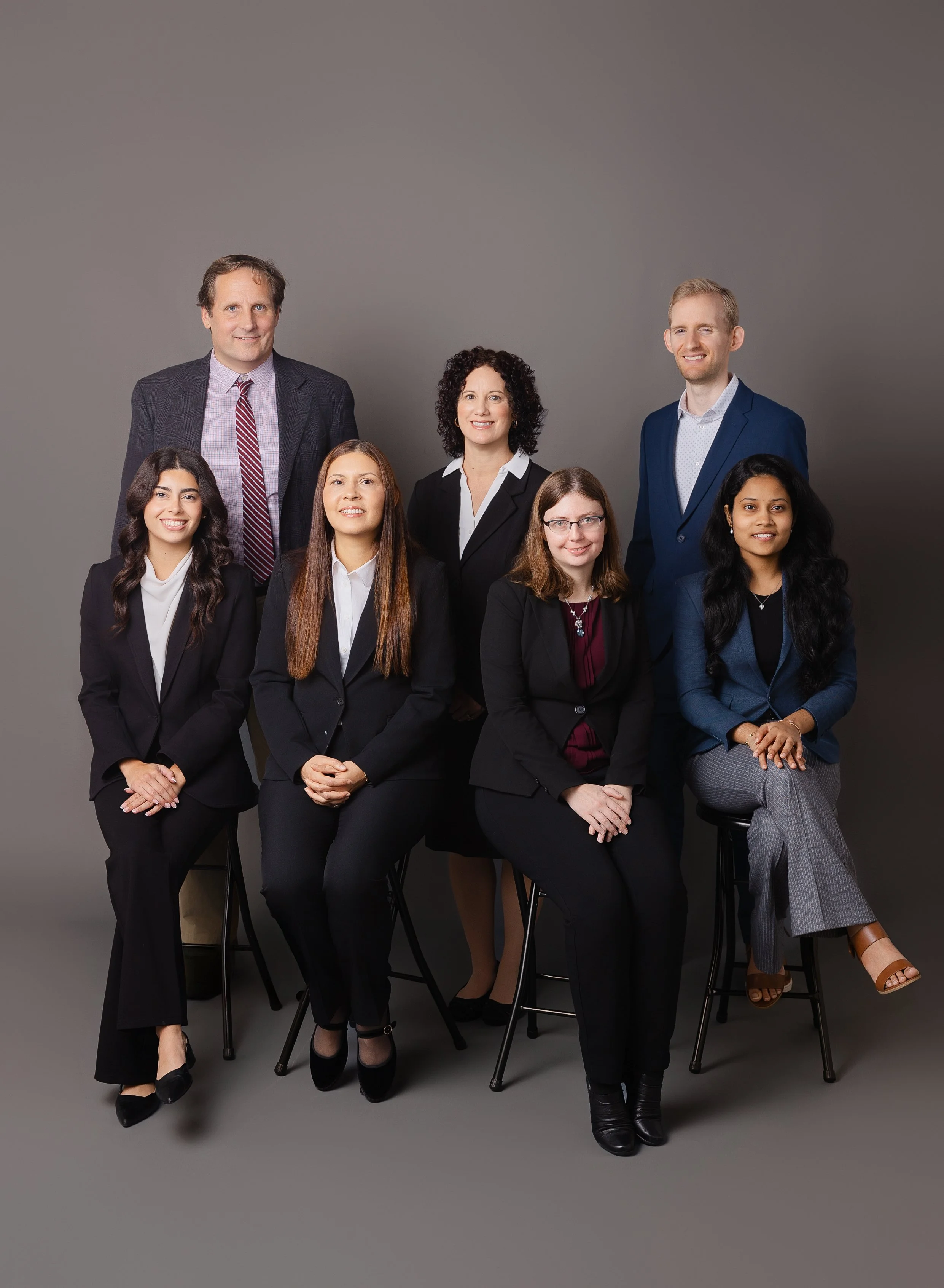 Group portrait of seven professionally dressed diverse adults, arranged in two rows against a gray background.