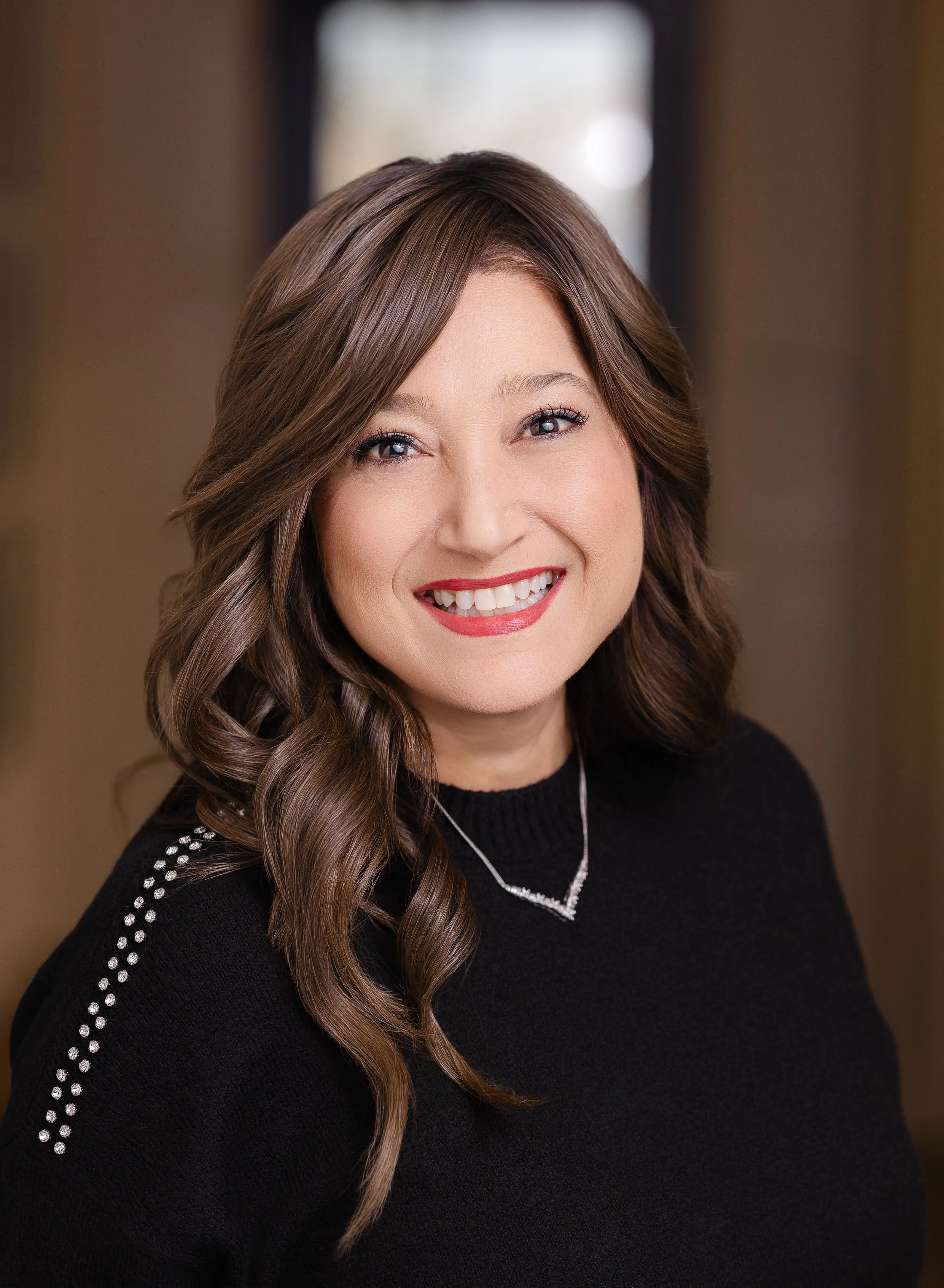 A woman with shoulder-length brown hair styled in loose curls, wearing a black sweater with rhinestone details on the shoulders, smiling with white teeth, in an indoor setting with a blurred window in the background.