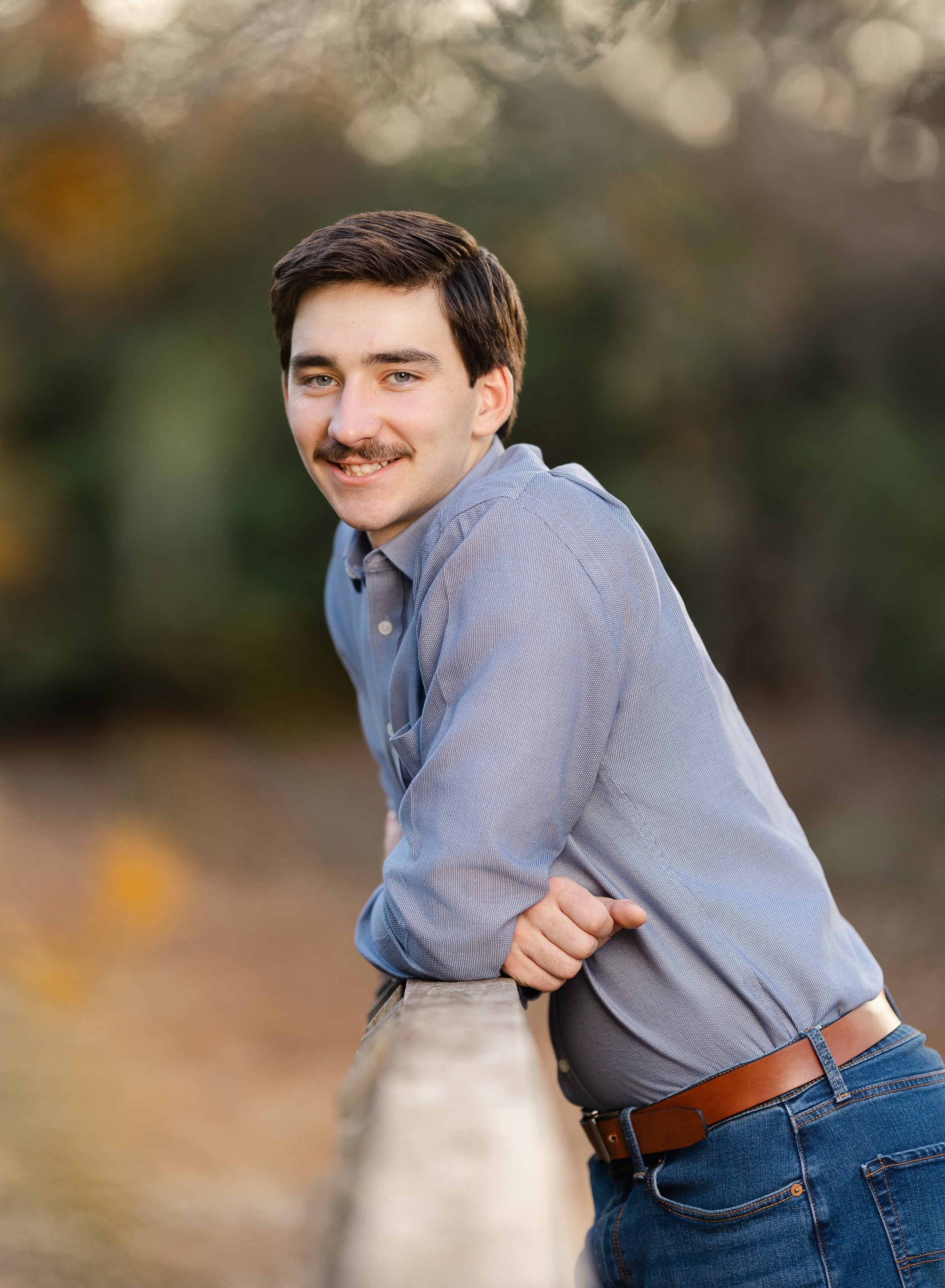 A young man with dark hair and a mustache smiling outdoors, leaning on a wooden railing with autumn foliage in the background.