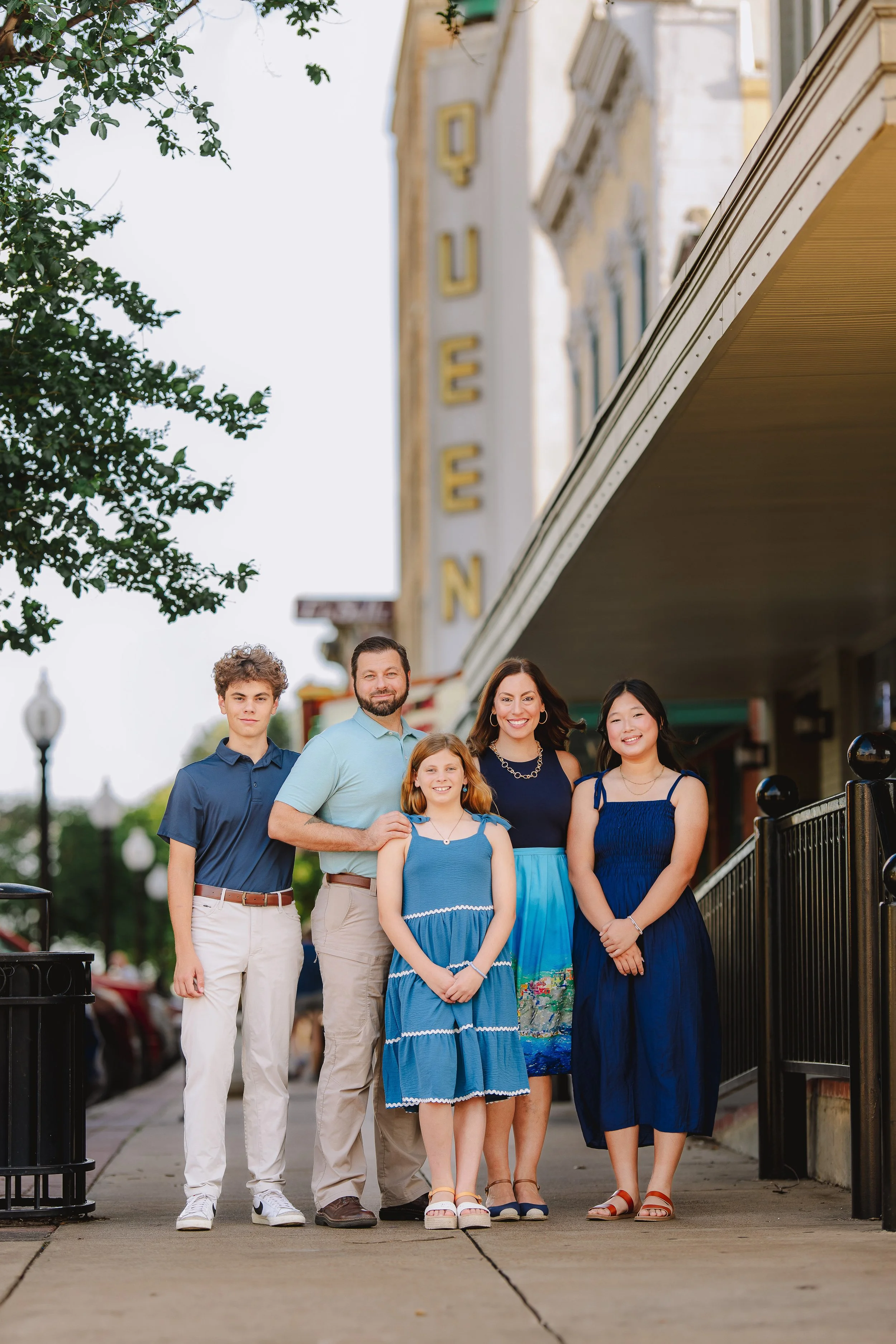 A family of five standing on a sidewalk in front of a building with a vertical sign that reads 'QUEEN.' The family members are smiling and dressed in blue and casual summer clothing.