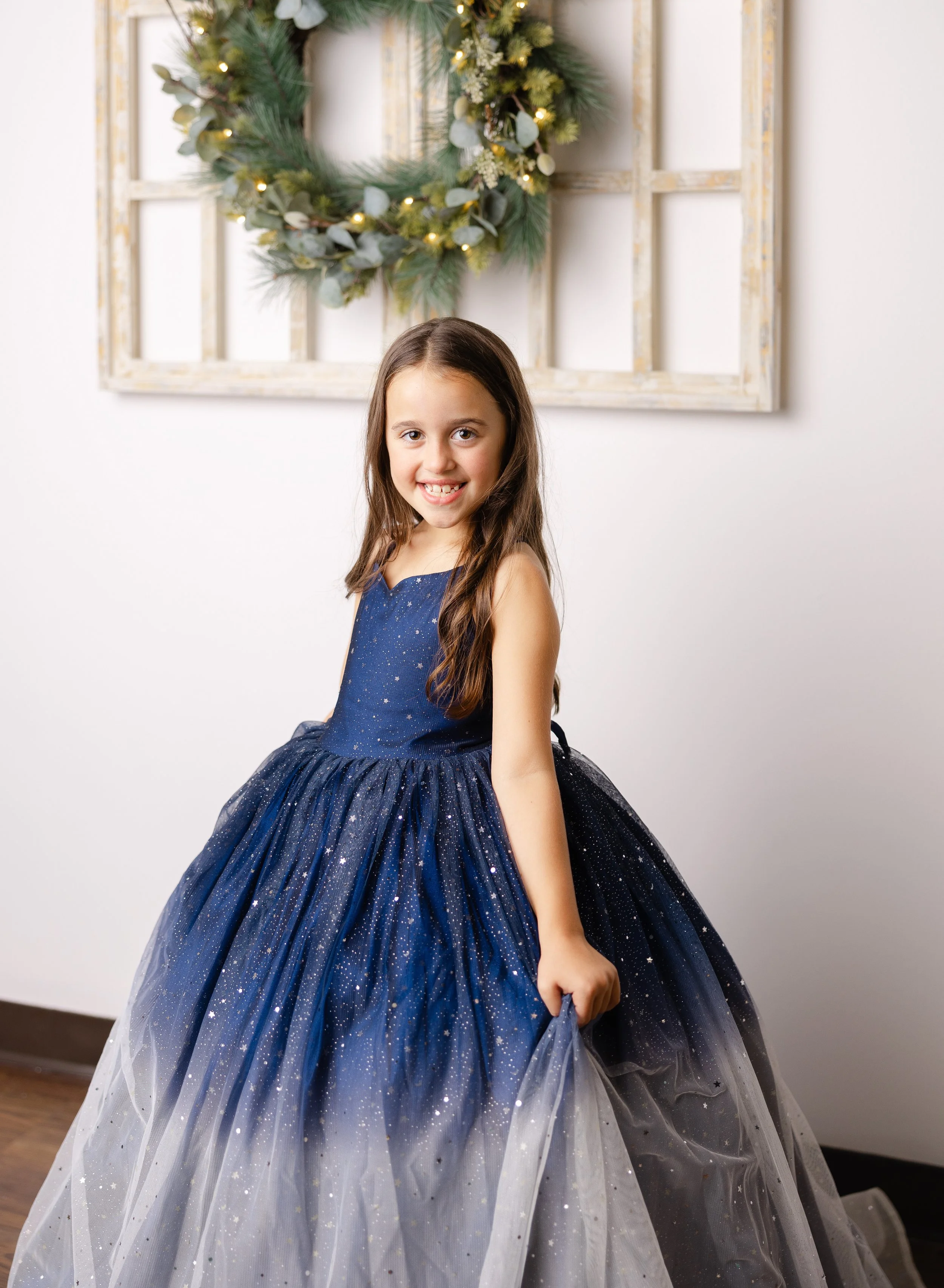 A young girl in a blue couture gown with sparkles, posing by a Christmas wreath and an antique window.