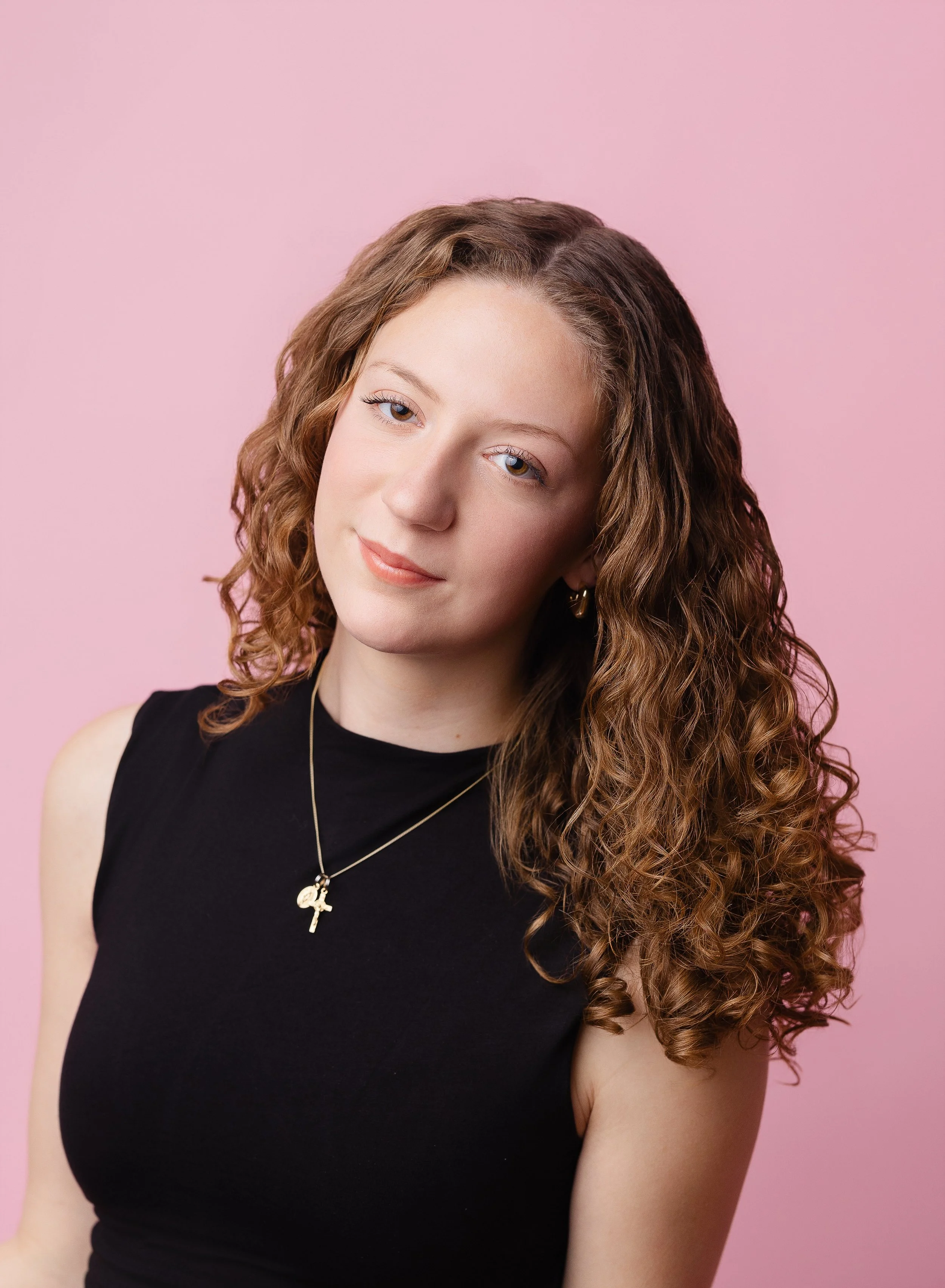 A young woman with wavy, shoulder-length brown hair and blue eyes, wearing a black sleeveless top and gold jewelry, sitting in front of a pink background.