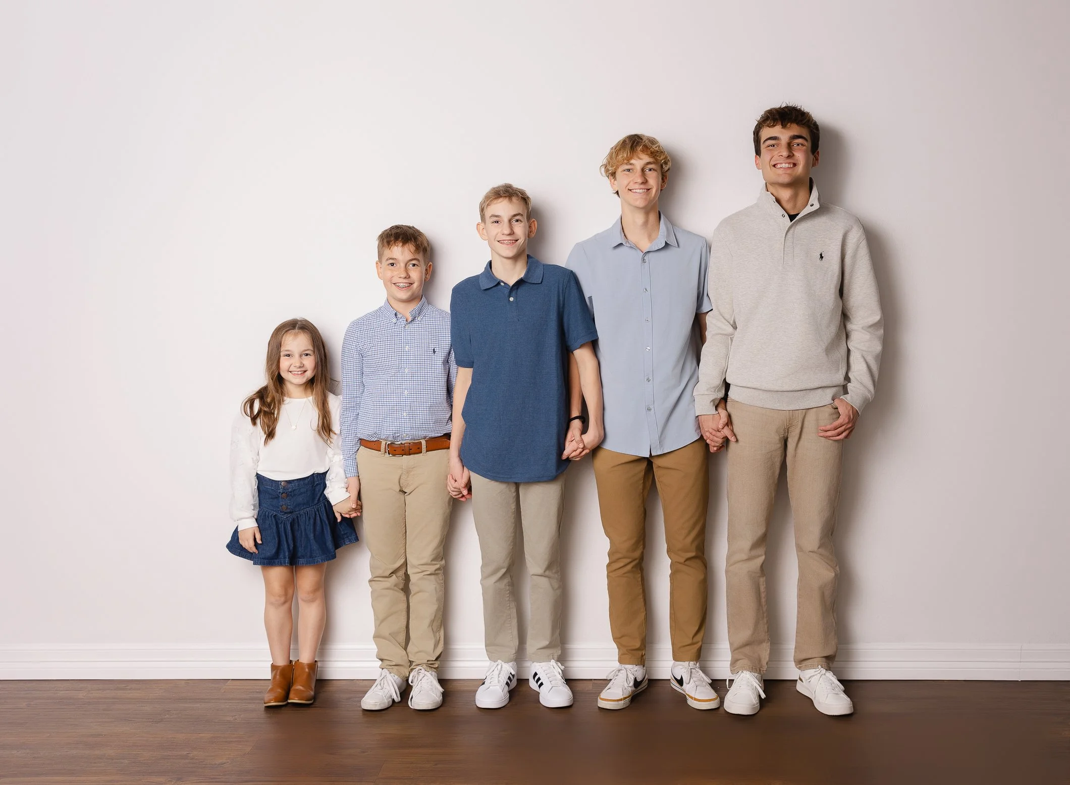 A group of six children standing in a line against a plain white wall, holding hands, smiling, dressed casually in light-colored clothing and sneakers.