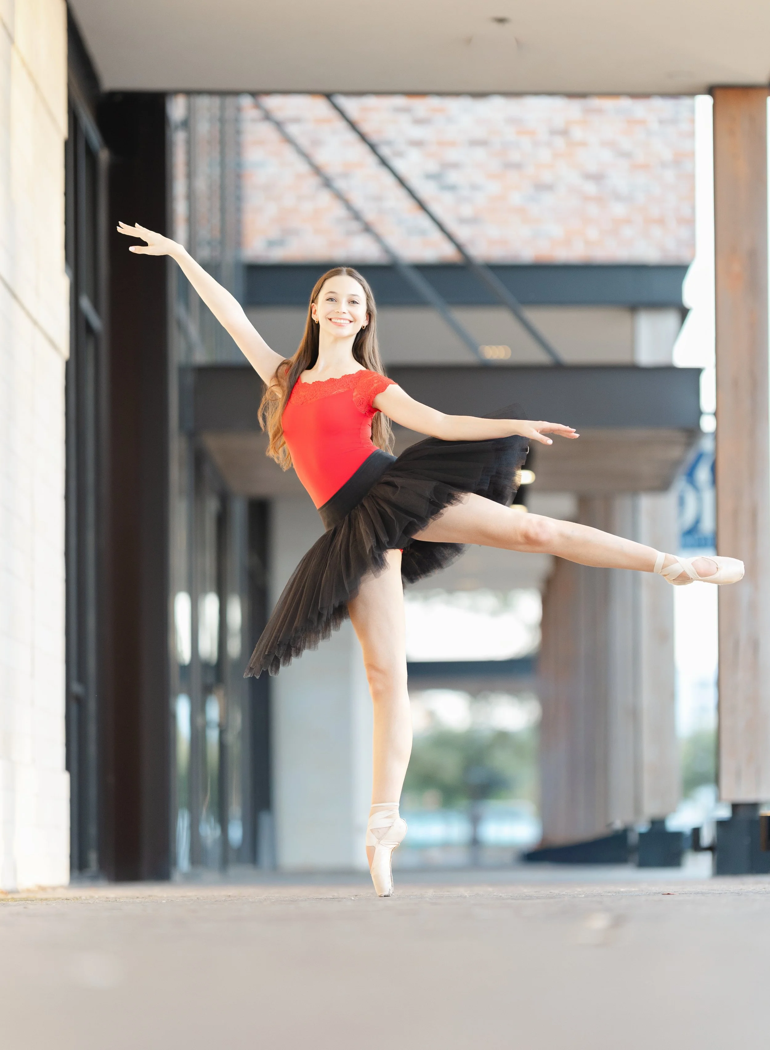 Ballet dancer in red top and black tutu practicing a pose on pointe shoes in an indoor space with modern architecture.