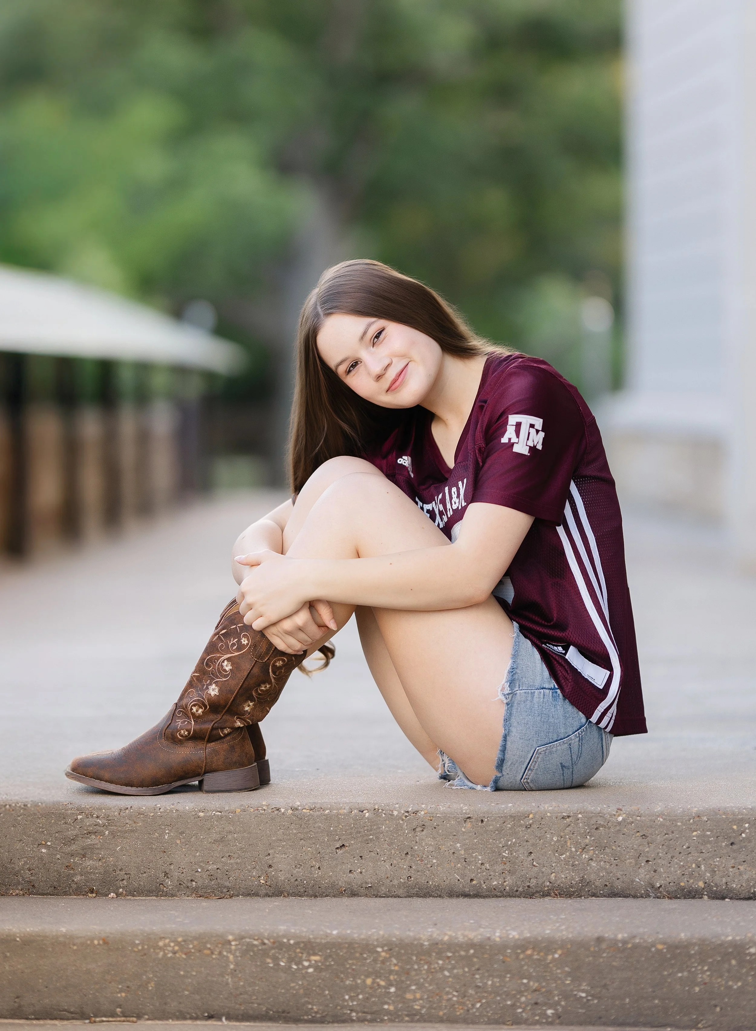A young girl with long brown hair sitting on the ground outside, smiling with her arms wrapped around her knees, wearing a maroon sports jersey, distressed denim shorts, and brown cowboy boots.