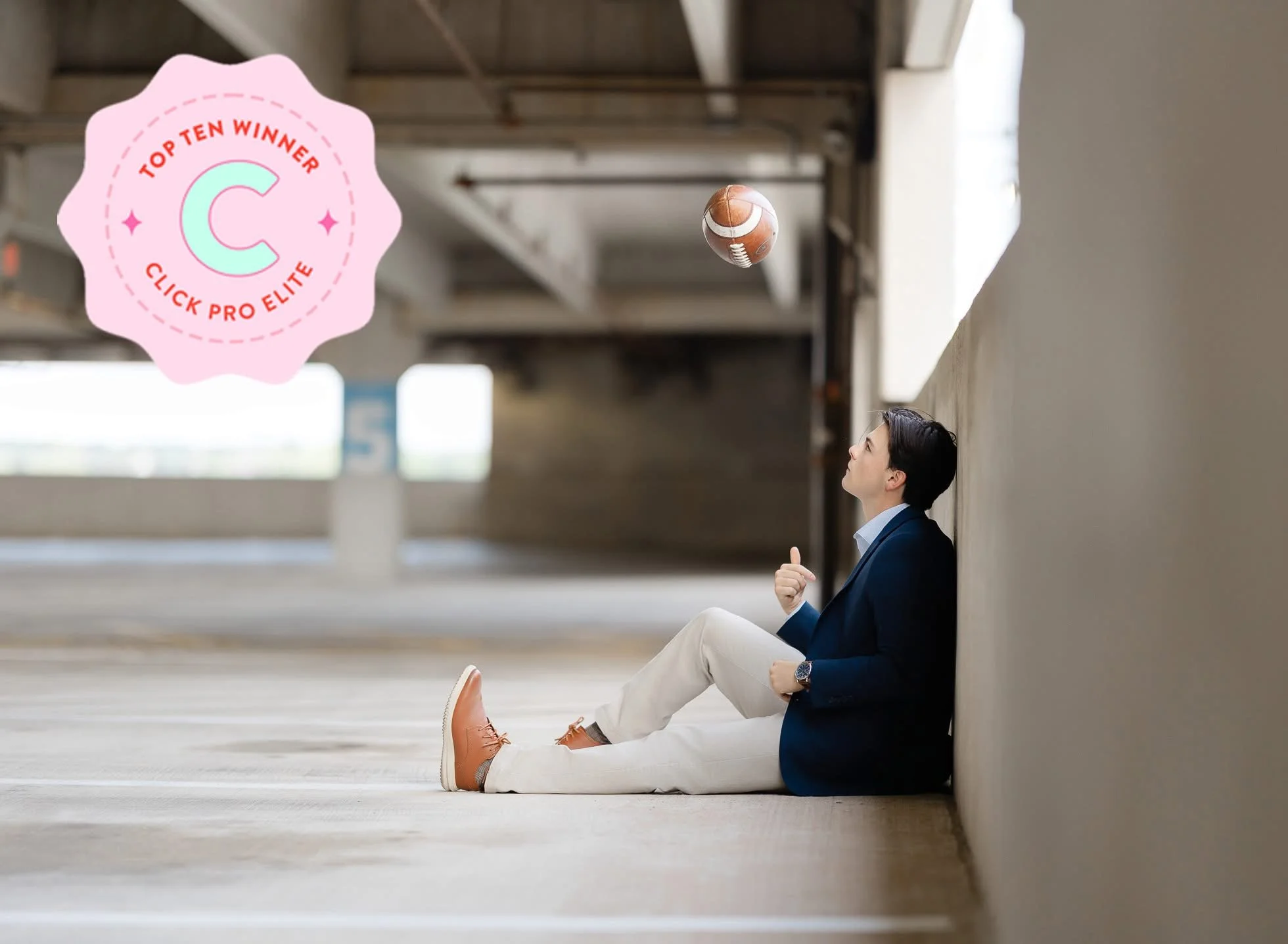 A man sitting on the ground with his back against a wall beneath a bridge or overpass, playing with a football. The man is wearing a dark blazer, light-colored pants, and shoes. There is a pink badge in the top left corner that reads "Top Ten Winner"