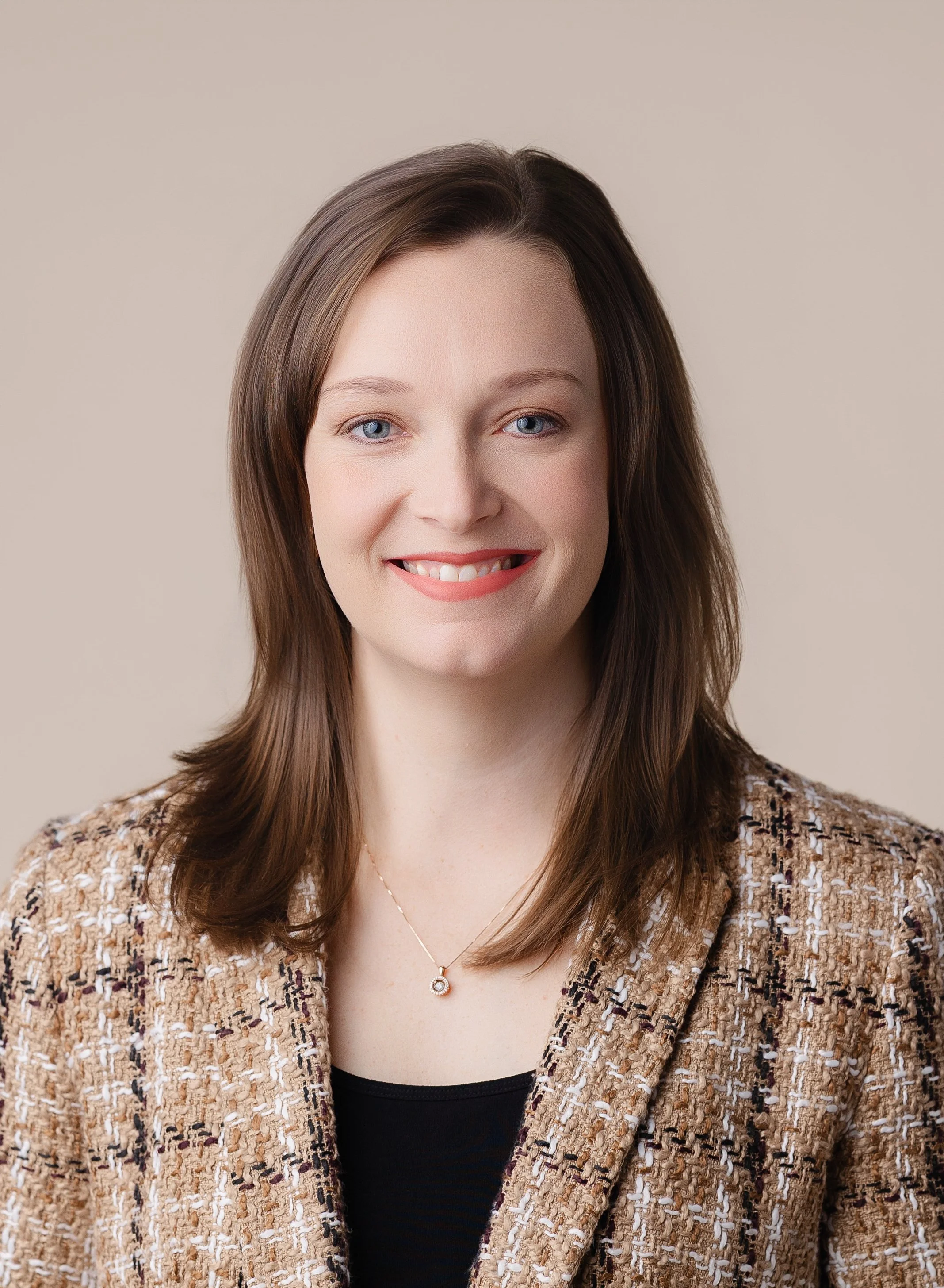 A woman with shoulder-length brown hair, blue eyes, and wearing a patterned beige blazer with a black top underneath, smiling against a neutral beige background.