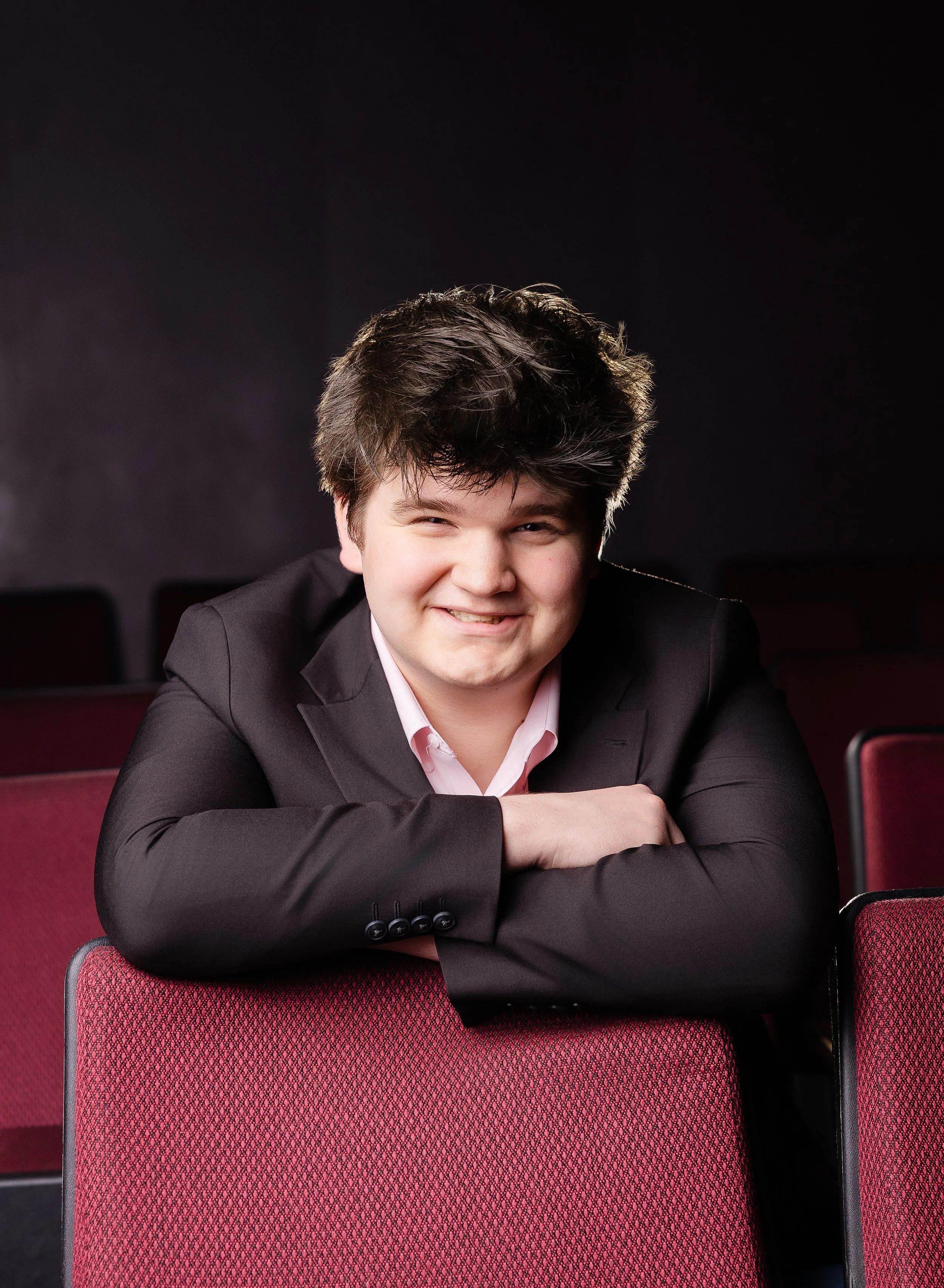 A young man with dark, curly hair smiling, wearing a black suit jacket and pink dress shirt, sitting in an auditorium with red cushioned seats.