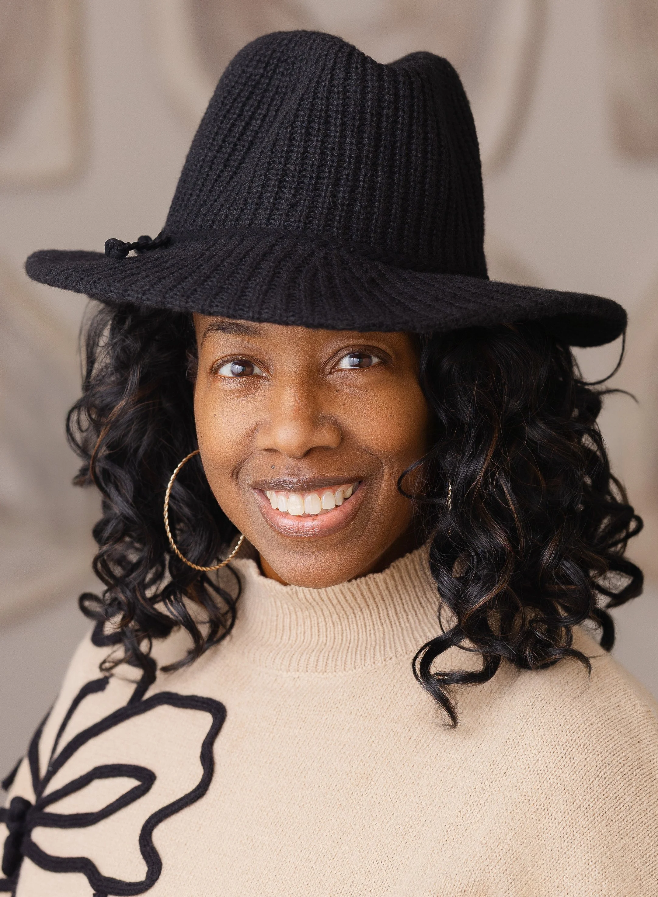 A woman wearing a beige sweater with black floral embroidery, large hoop earrings, and a black knit wide-brimmed hat, smiling at the camera.