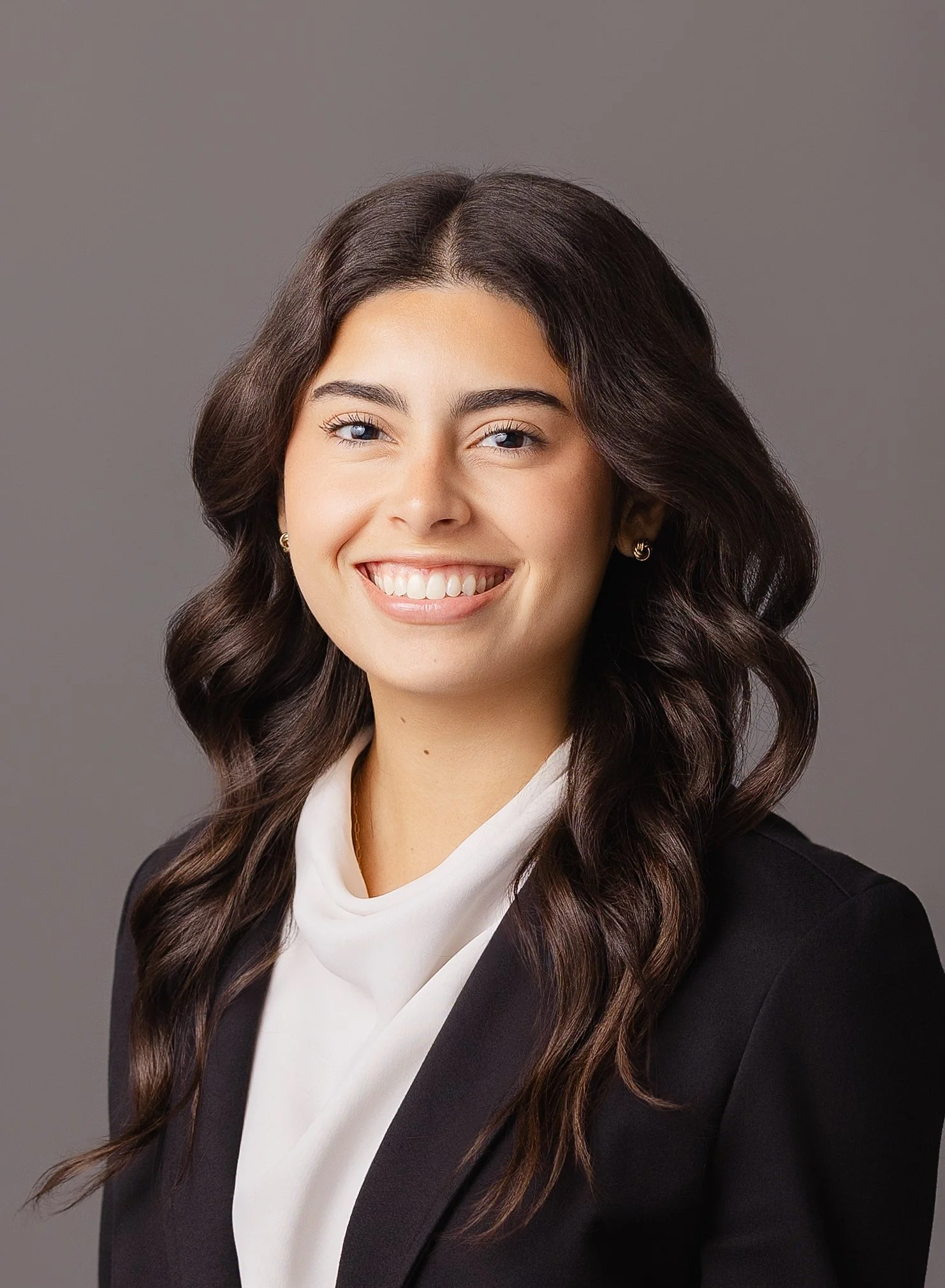 A woman with long, wavy dark brown hair smiling, wearing a black blazer and white blouse, against a gray background.